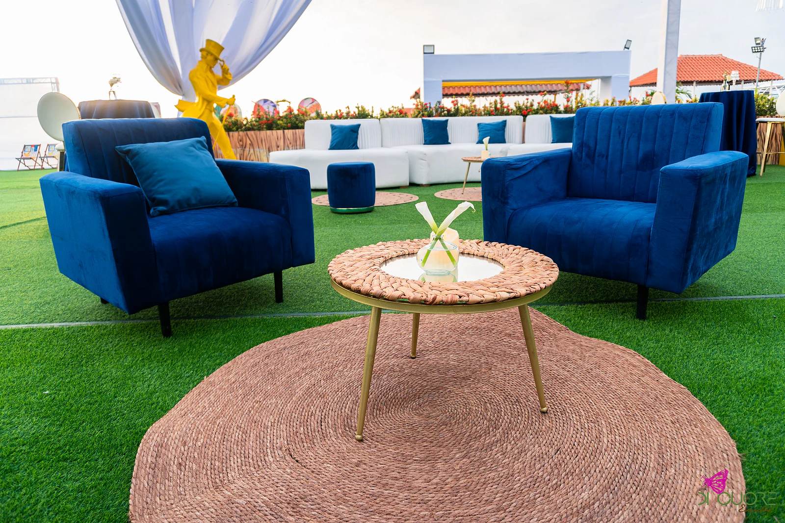 Duas poltronas de veludo azul e uma mesa baixa sobre um tapete de corda, na relva do lounge exterior