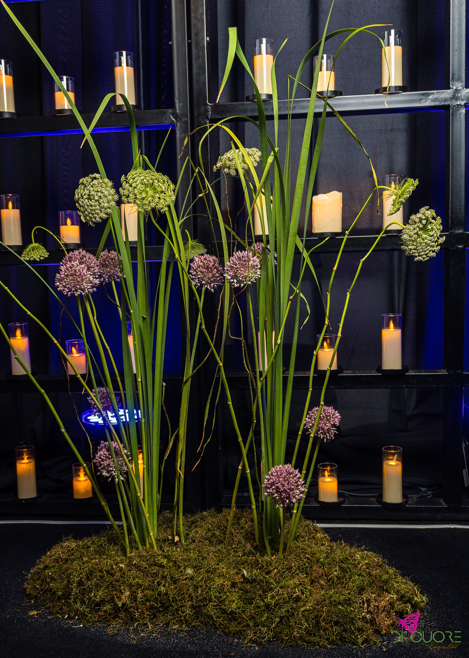 Ilha de musgo com alhos ornamentais, umbelas verdes e capim alto, diante de prateleiras de velas