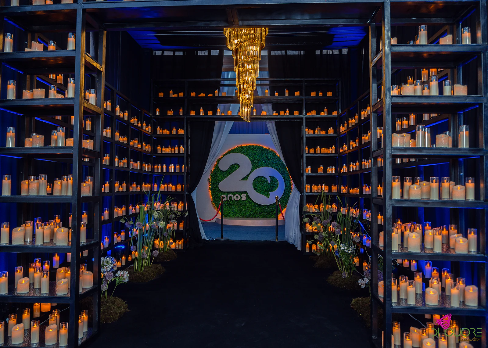 Corredor de prateleiras de aço cheias de velas, com um lustre dourado e o logótipo “20 anos” num círculo de folhagem ao fundo