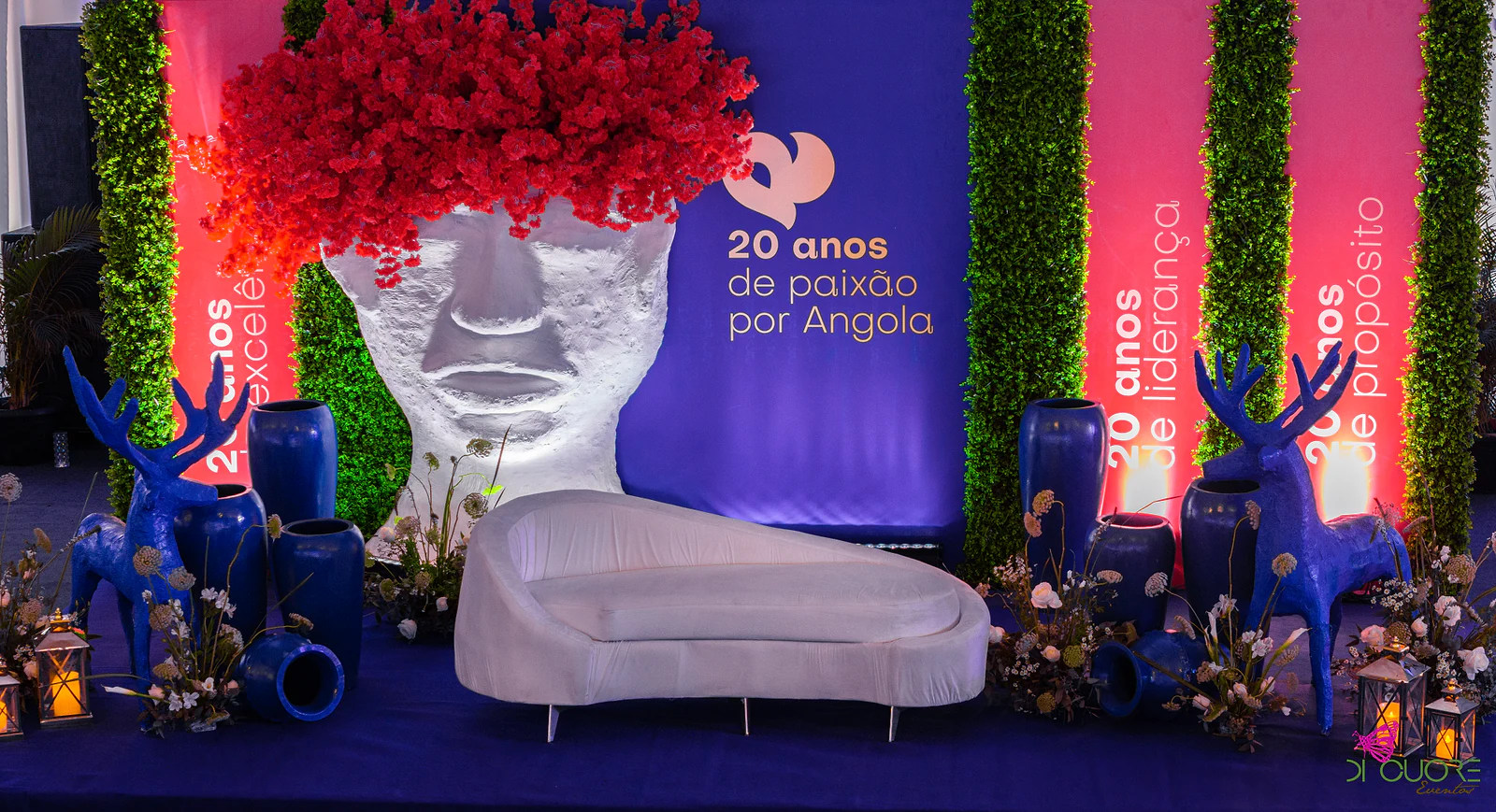 Palco com a escultura de rosto de cabeleira vermelha, sofá branco curvo, veados e jarros azuis, e o painel “20 anos de paixão por Angola”