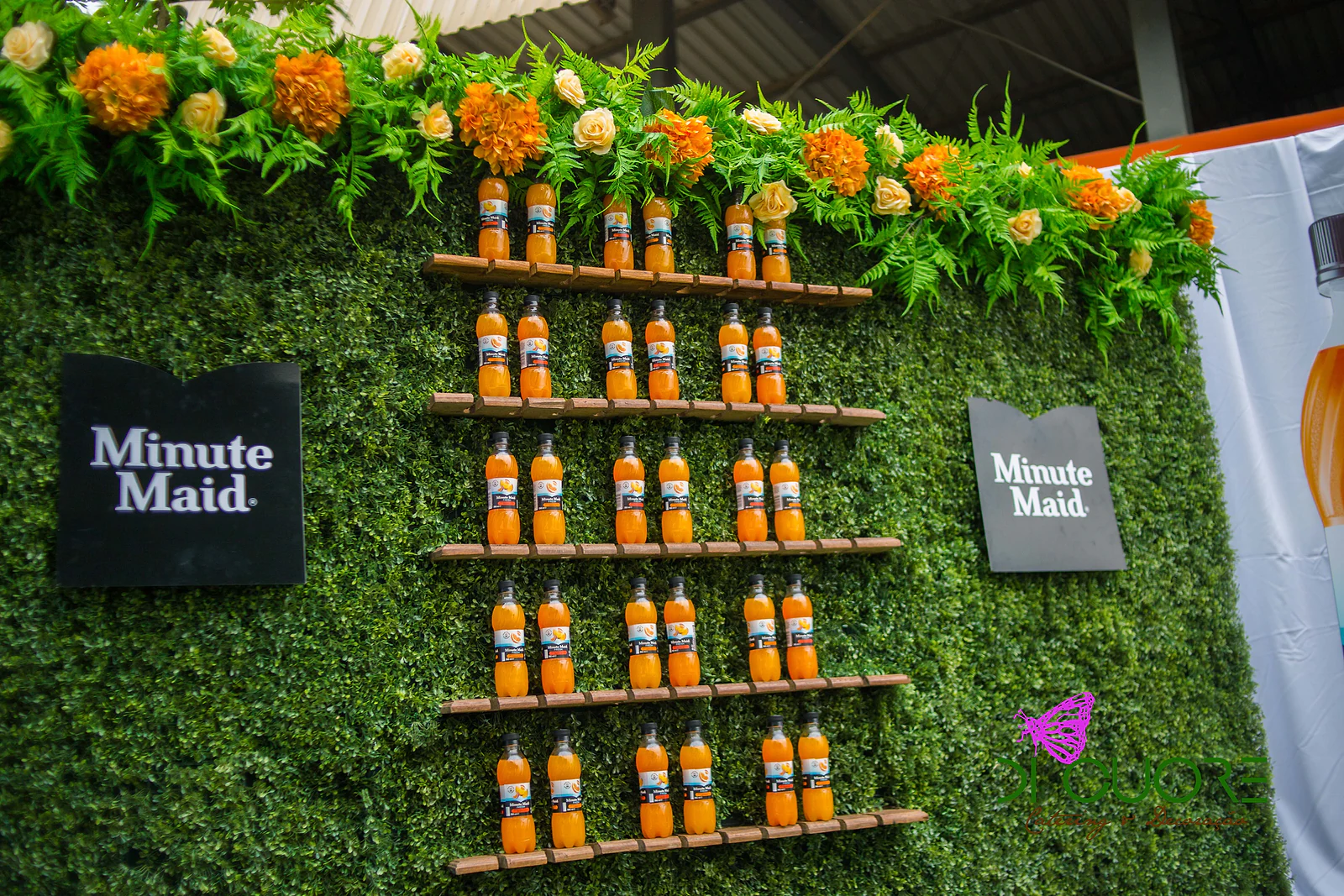 Muro de buxo com prateleiras de madeira e garrafas de sumo de laranja, coroado por flores cor de laranja e fetos