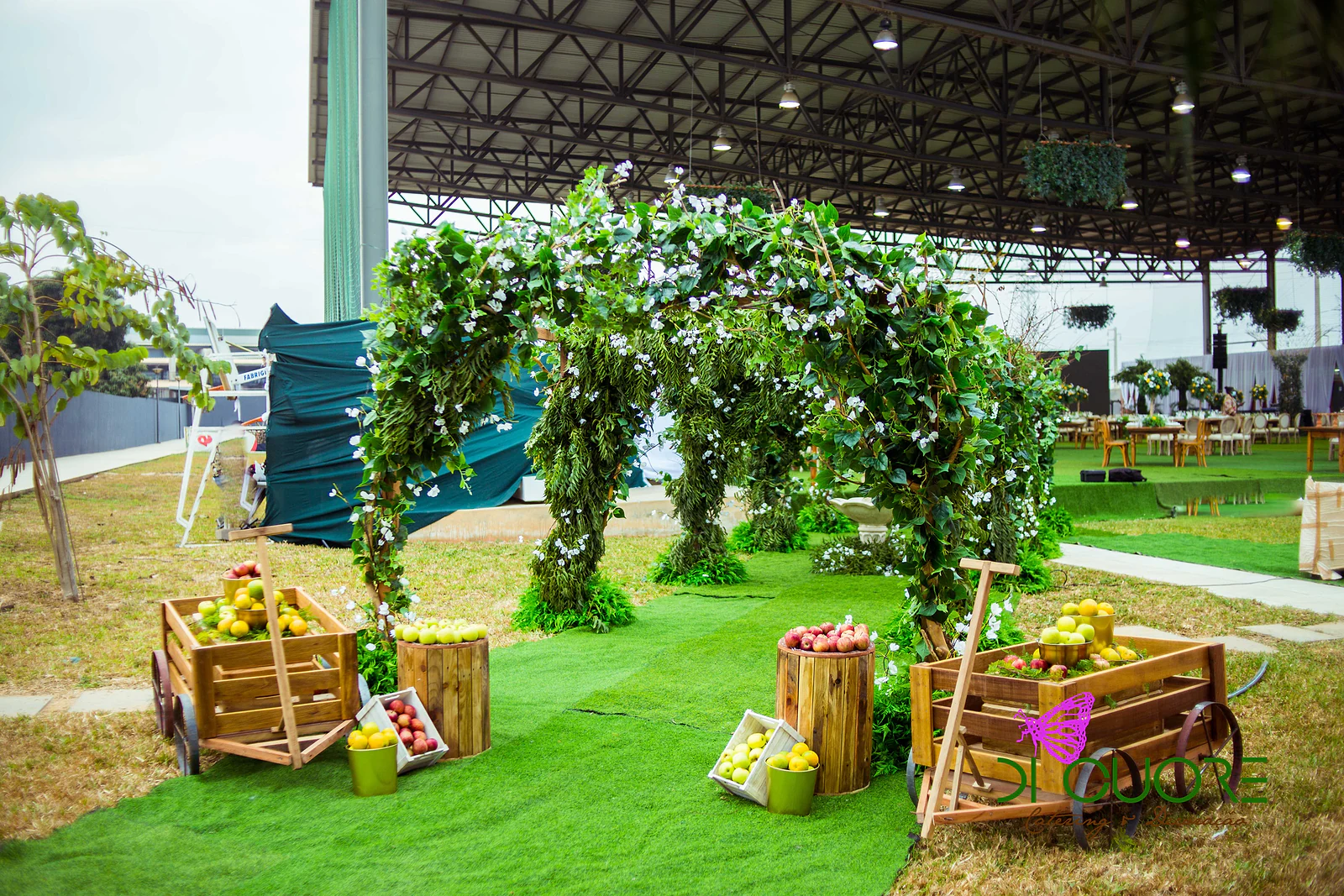 Arcos de folhagem e pequenas flores brancas sobre o relvado, com carrinhos de madeira e barris cheios de maçãs e limões