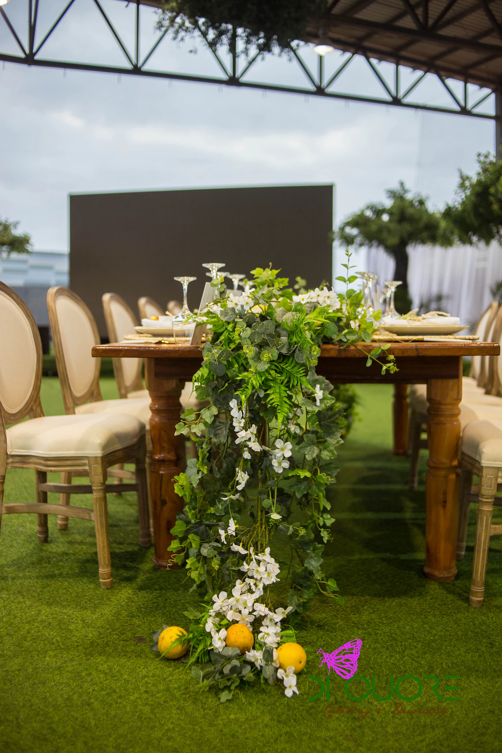 Ponta de mesa de madeira com folhagem e flores brancas a cair até ao chão, entre cadeiras de costas redondas, com limões na relva