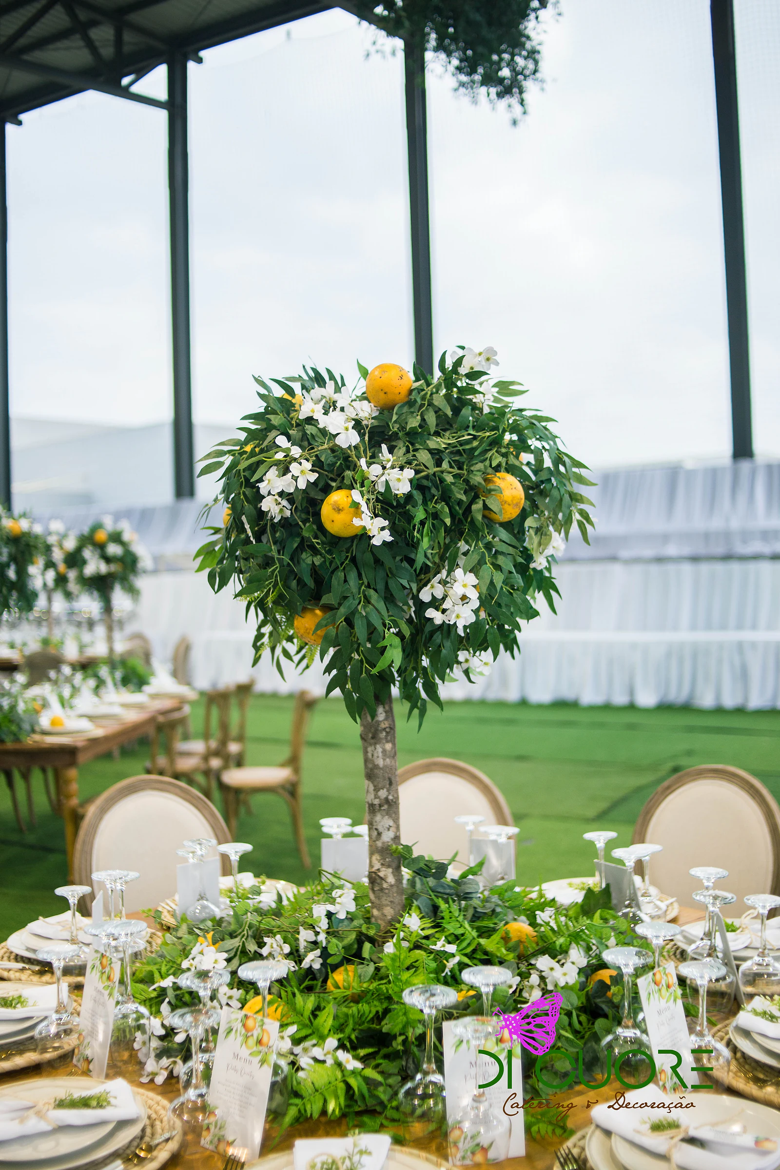 Limoeiro decorativo ao centro de uma mesa posta, com folhagem, flores brancas, limões e ementas ilustradas