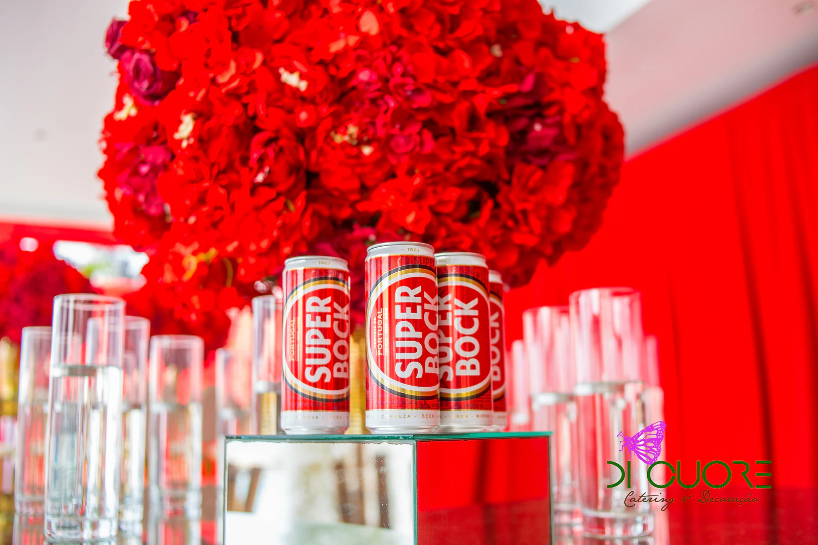 Esfera de flores vermelhas sobre um pedestal de espelho, com latas de Super Bock e copos altos de vidro diante de um cortinado vermelho