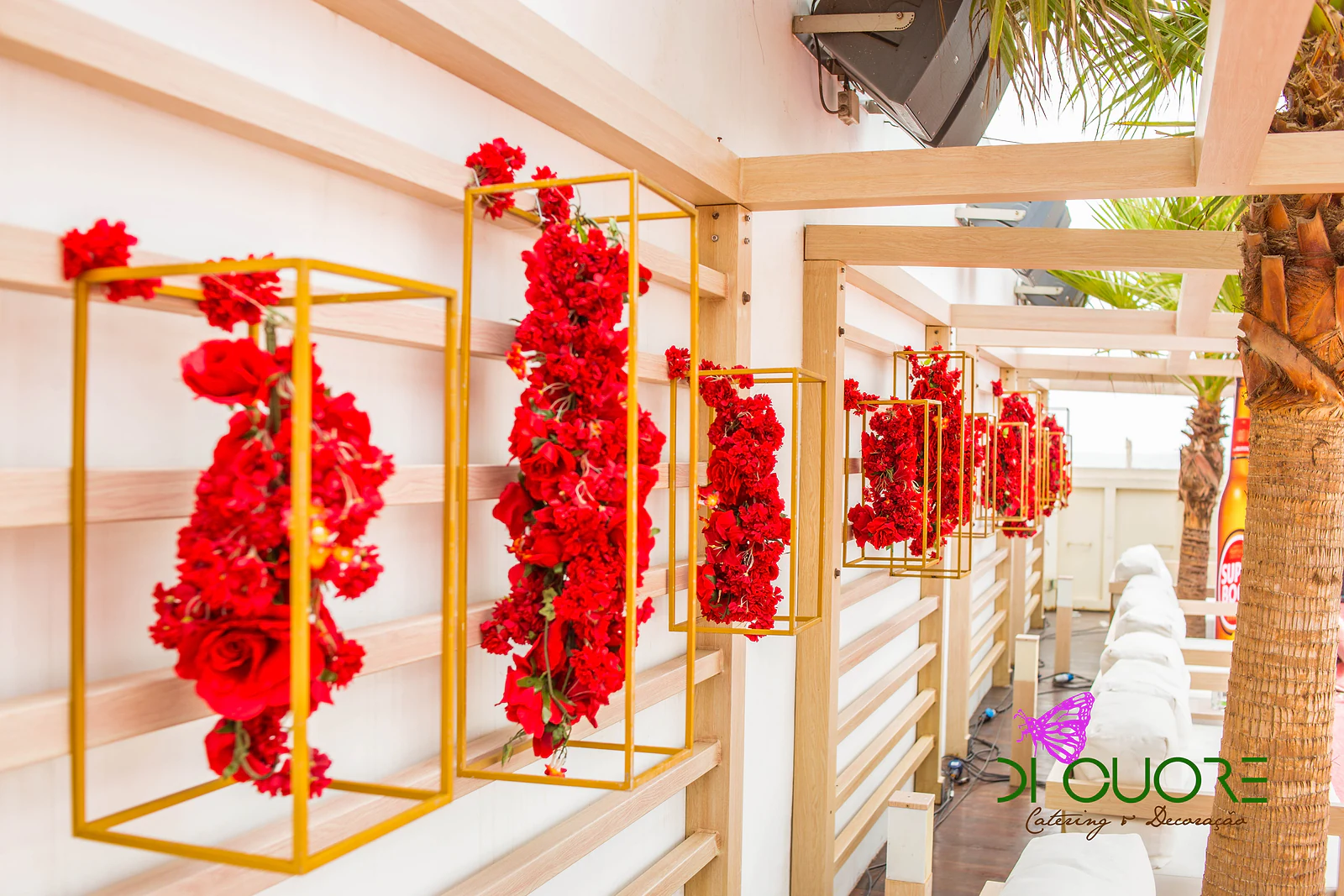 Ramos de flores vermelhas dentro de molduras douradas, presas a uma parede de ripas claras, junto a uma palmeira