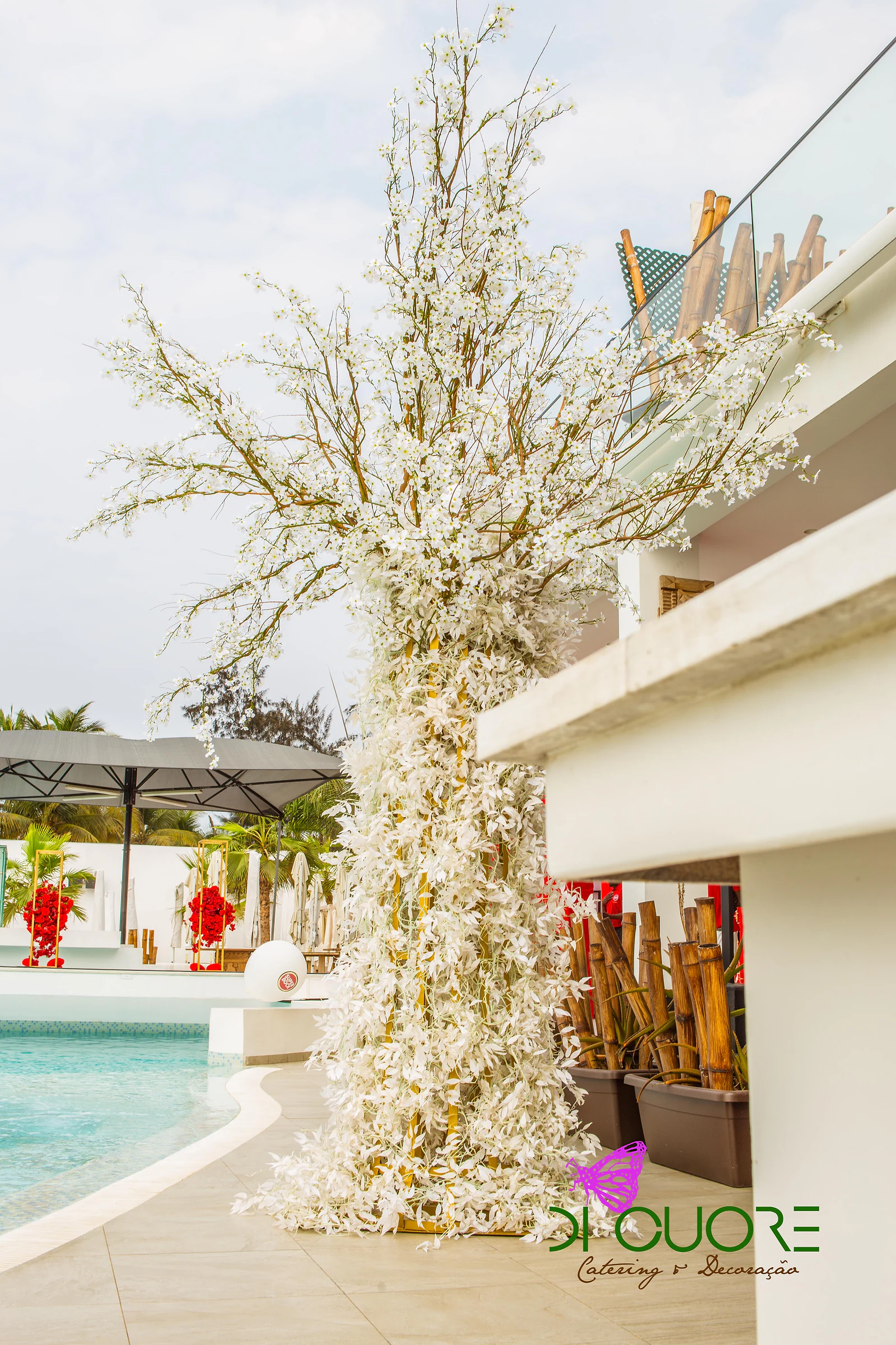 Árvore de ramos cobertos de flores brancas junto a uma piscina de água turquesa, com bambus em vasos ao lado