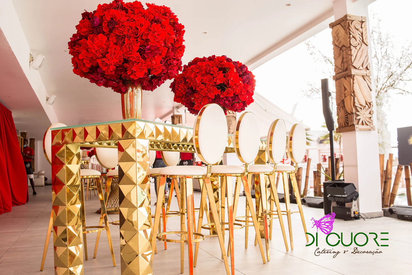Mesa alta dourada de relevo facetado, com grandes esferas de flores vermelhas e bancos altos brancos de armação dourada