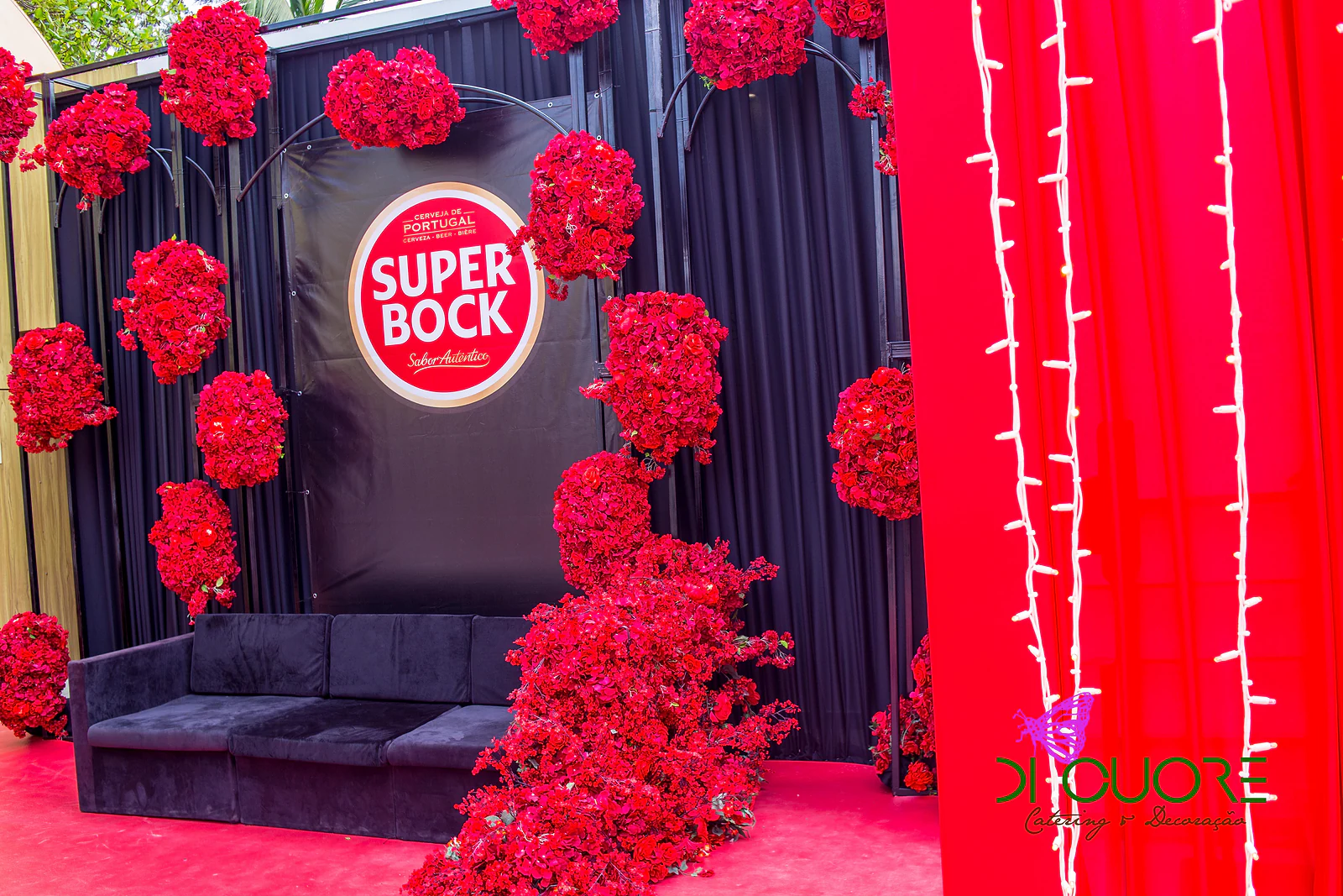Fundo fotográfico com o logótipo Super Bock sobre cortinado preto, com esferas e cascata de flores vermelhas e um sofá de veludo preto
