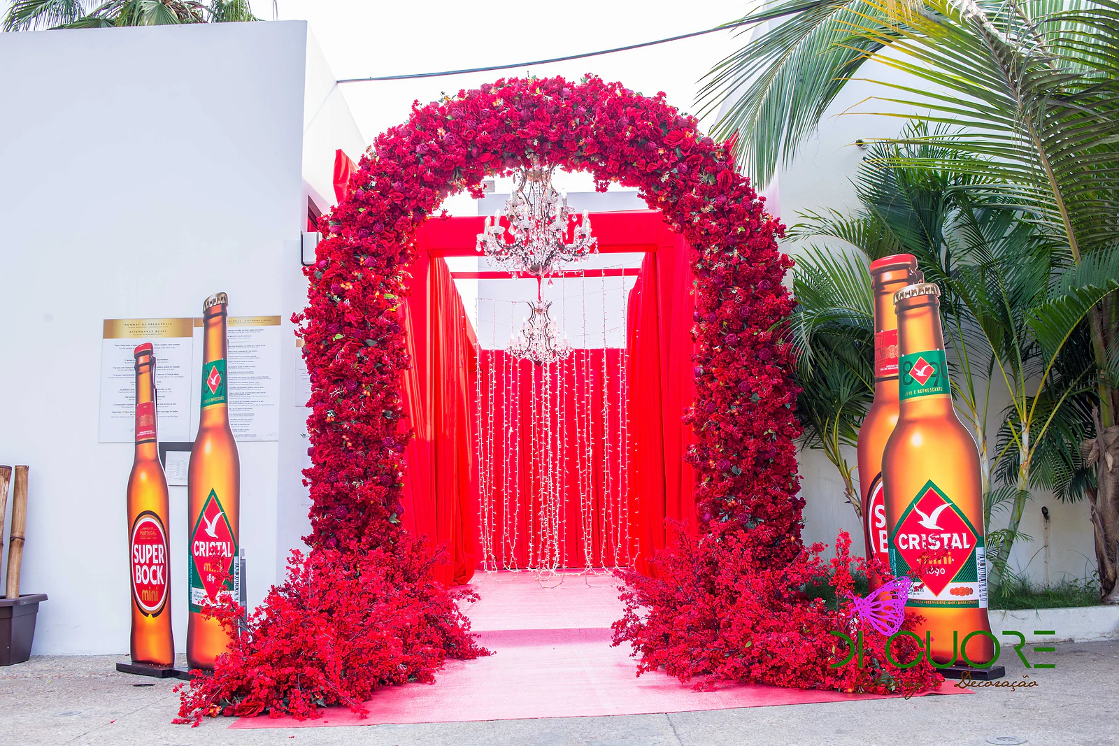 Arco de rosas vermelhas sobre uma passadeira rosa, com lustre de cristal e cortinados vermelhos ao fundo, entre palmeiras e garrafas gigantes de cerveja Cristal e Super Bock