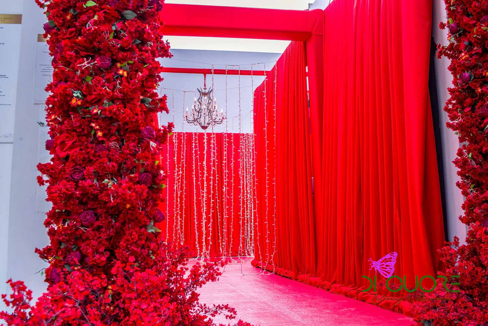 Coluna de rosas vermelhas junto a cortinados vermelhos e fios de luzes, com um lustre de cristal ao fundo do corredor