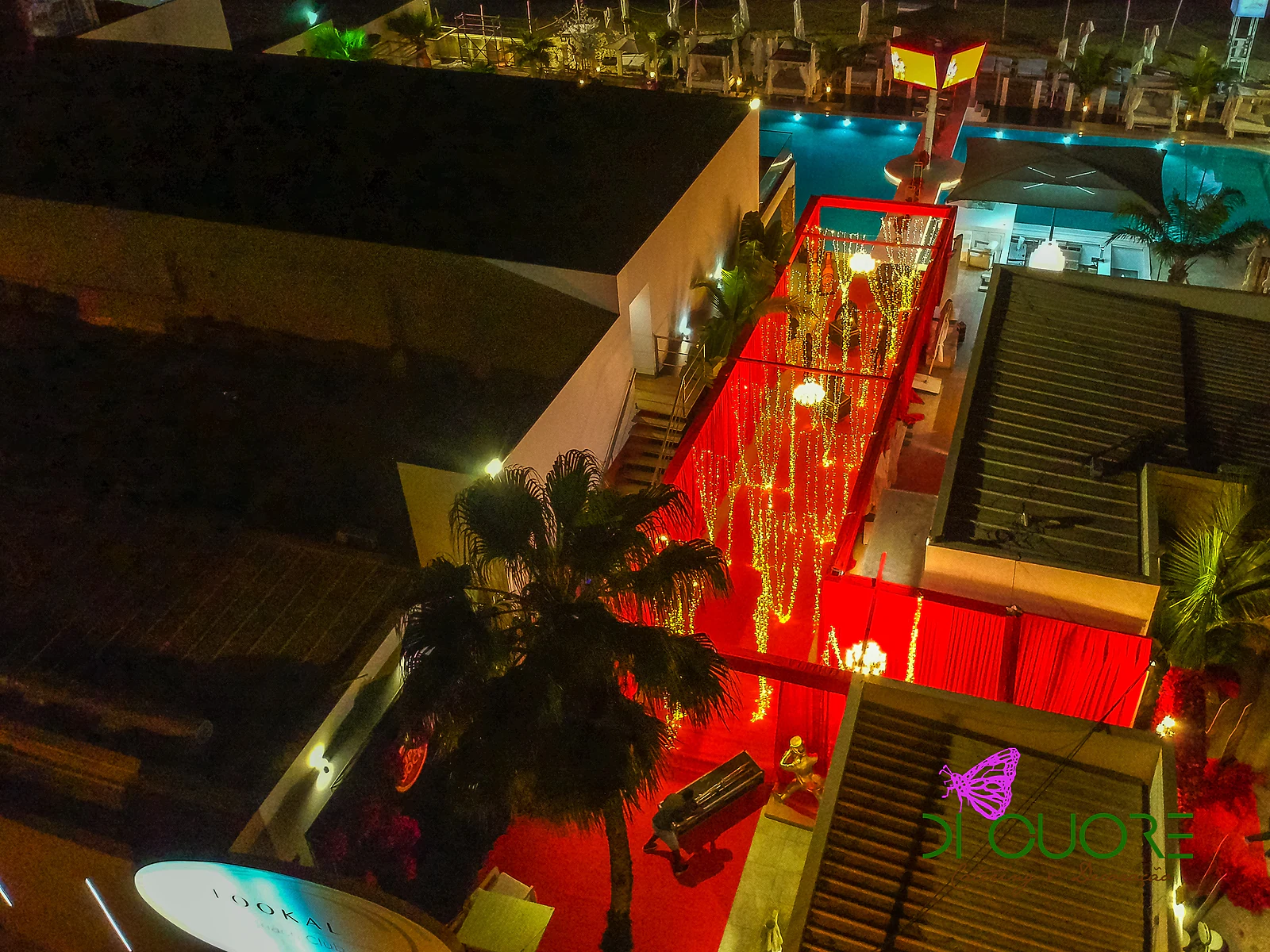 Vista de cima, à noite, do corredor de cortinados vermelhos e fios de luzes, junto à piscina iluminada