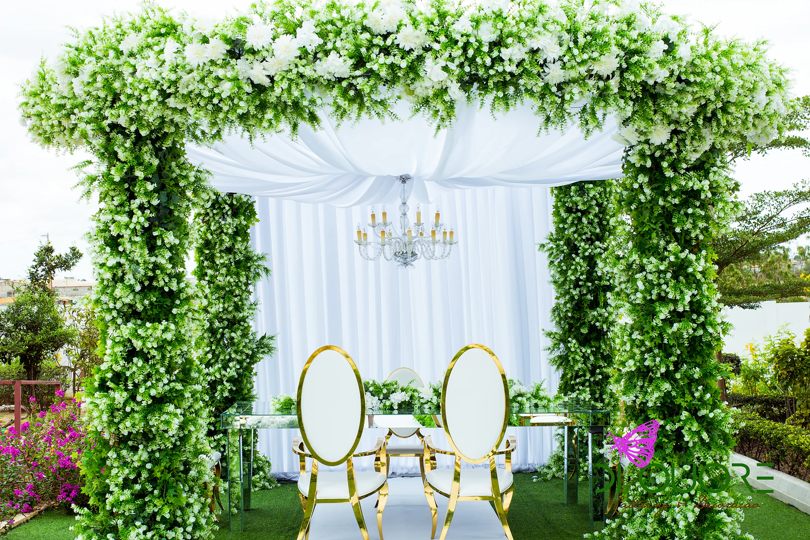 Pérgola revestida de folhagem e pequenas flores brancas, com cortinado branco, lustre de cristal e dois cadeirões de encosto oval dourado