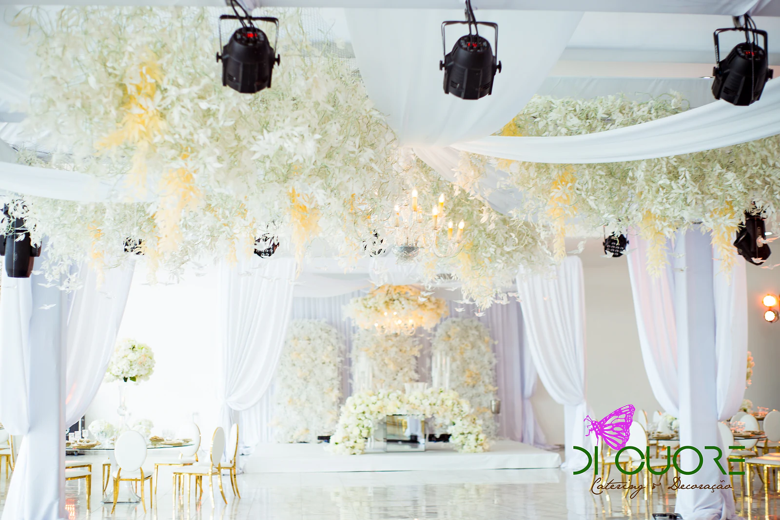 Salão de tecto coberto de flores brancas e creme suspensas, com cortinados brancos, projectores e o arco florido ao fundo