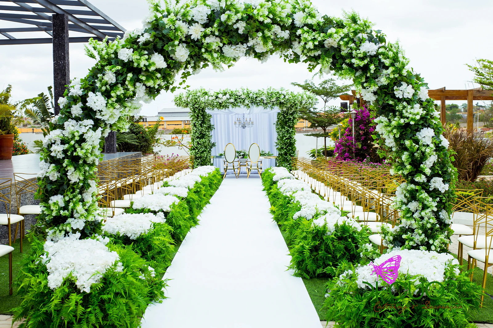 Passadeira branca entre canteiros de fetos e hortênsias brancas, sob um arco de flores brancas e folhagem, com cadeiras douradas e a pérgola ao fundo