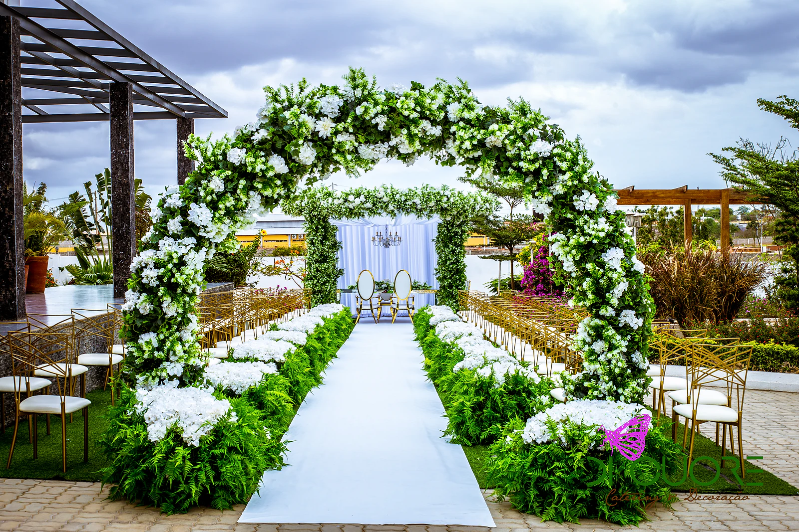 Arco de flores brancas e folhagem sobre uma passadeira branca, com cadeiras douradas dos lados e um altar com cortinados ao fundo