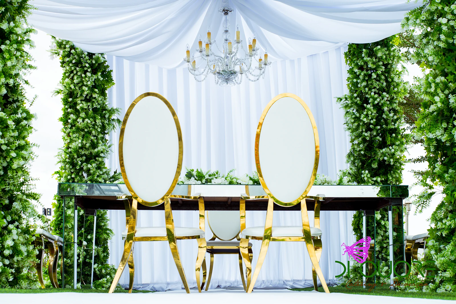 Dois cadeirões brancos de encosto oval e armação dourada diante de uma mesa escura, sob cortinado branco e lustre de cristal, entre colunas de folhagem