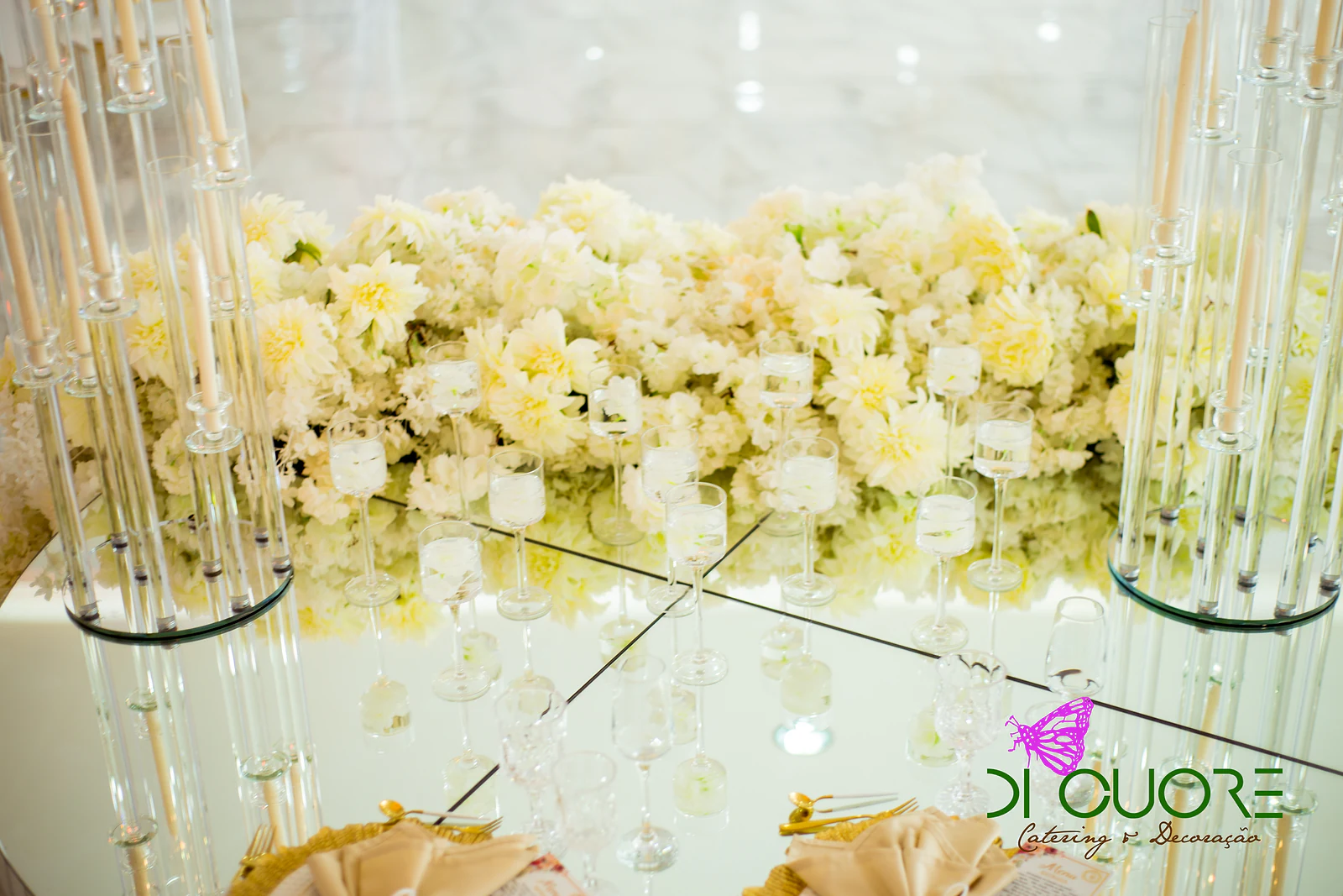 Fileira de flores creme sobre uma mesa de espelho, entre candelabros altos de vidro e pequenos castiçais