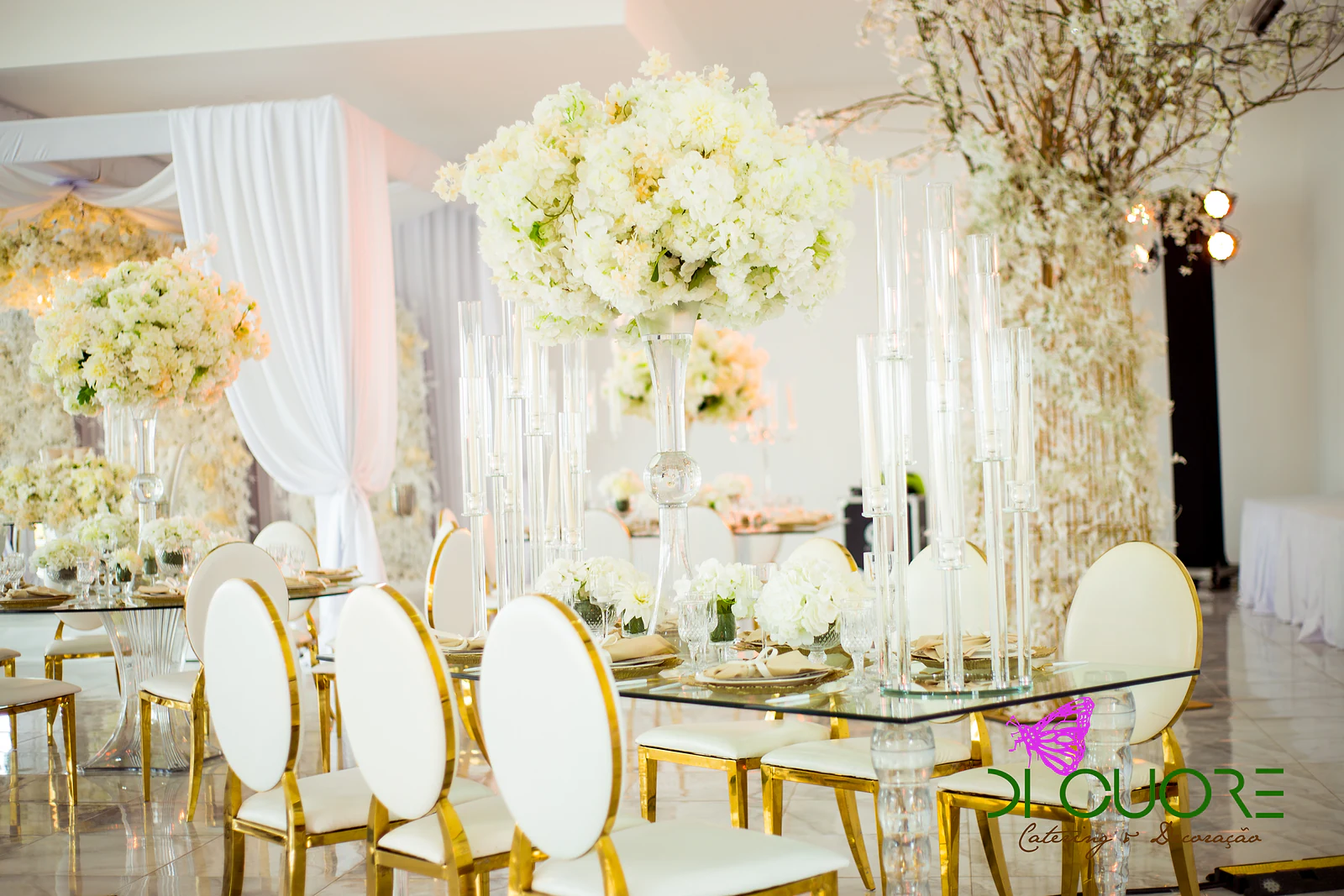 Mesa de vidro com ramos volumosos de flores creme, candelabros altos de vidro e cadeiras brancas de armação dourada