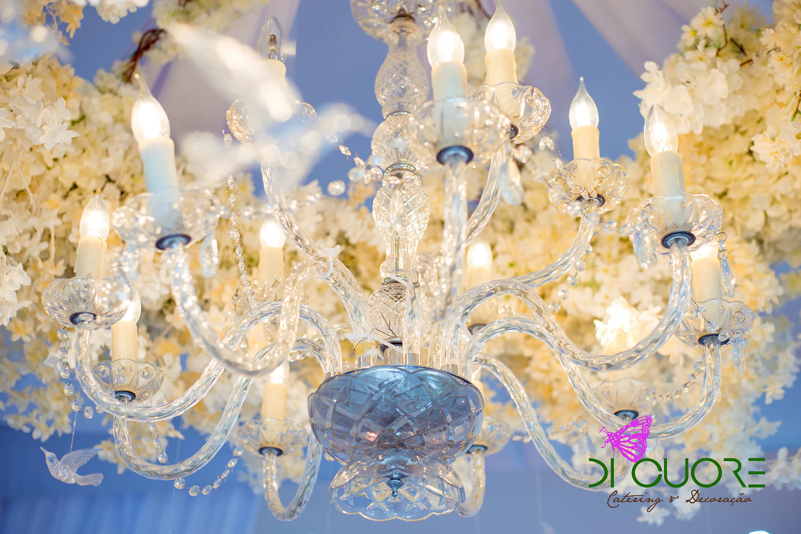 Lustre de cristal com velas acesas, entre flores brancas suspensas, sobre um fundo de luz azul