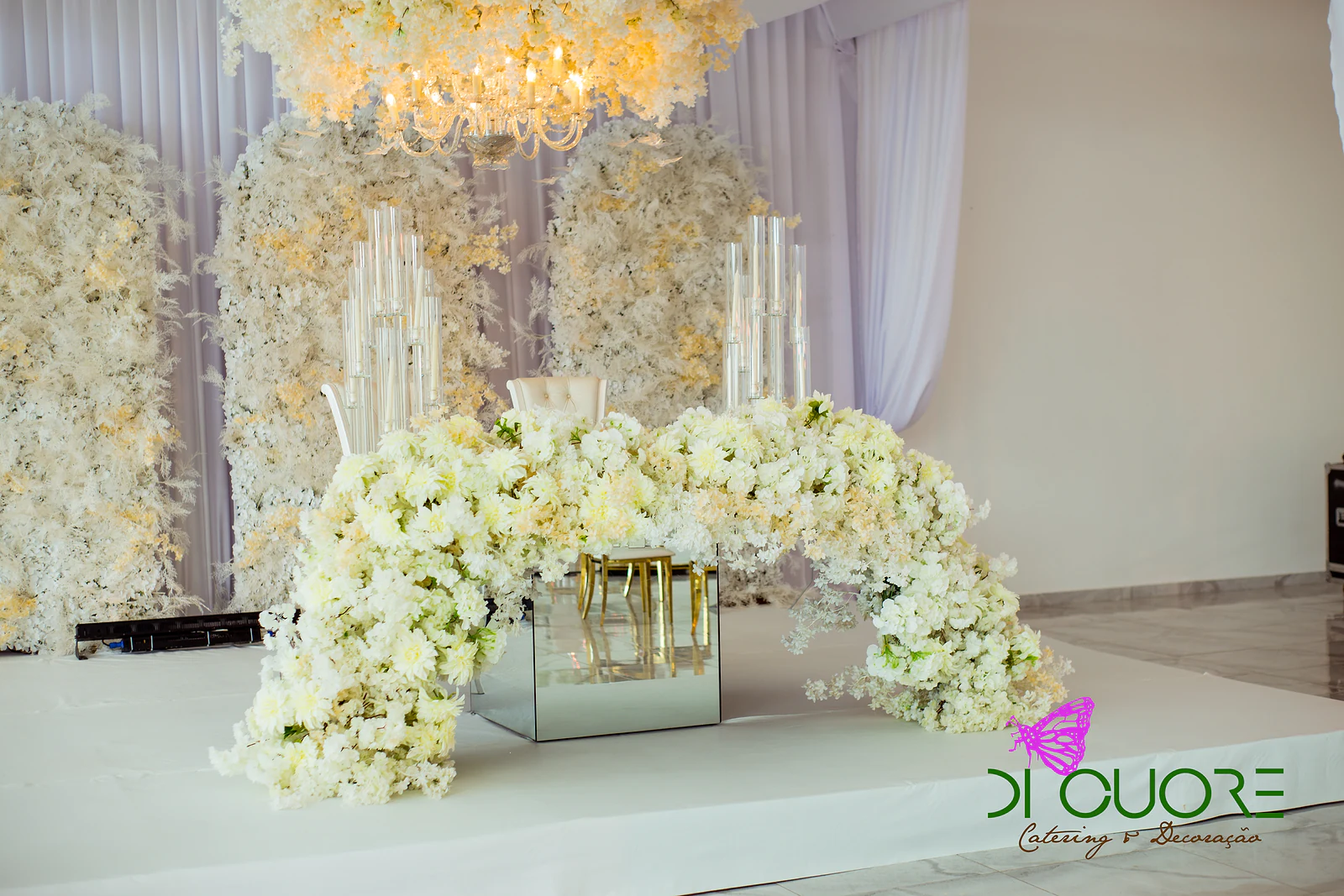 Arco de flores creme sobre um cubo de espelho, diante de paredes de flores brancas e candelabros de vidro, sob um lustre florido