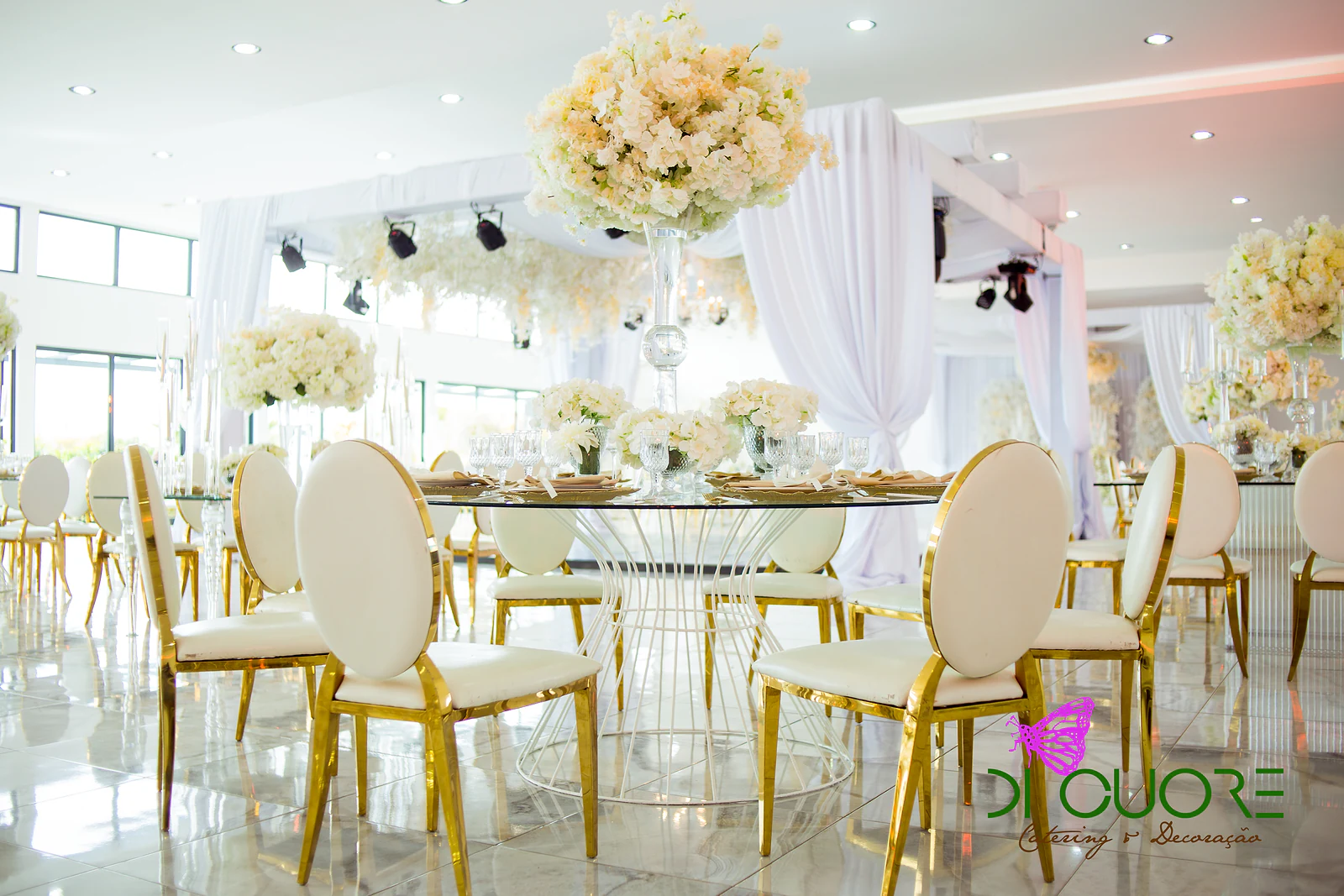 Mesa redonda de vidro com um grande ramo de flores creme, rodeada de cadeiras brancas de armação dourada, num salão luminoso