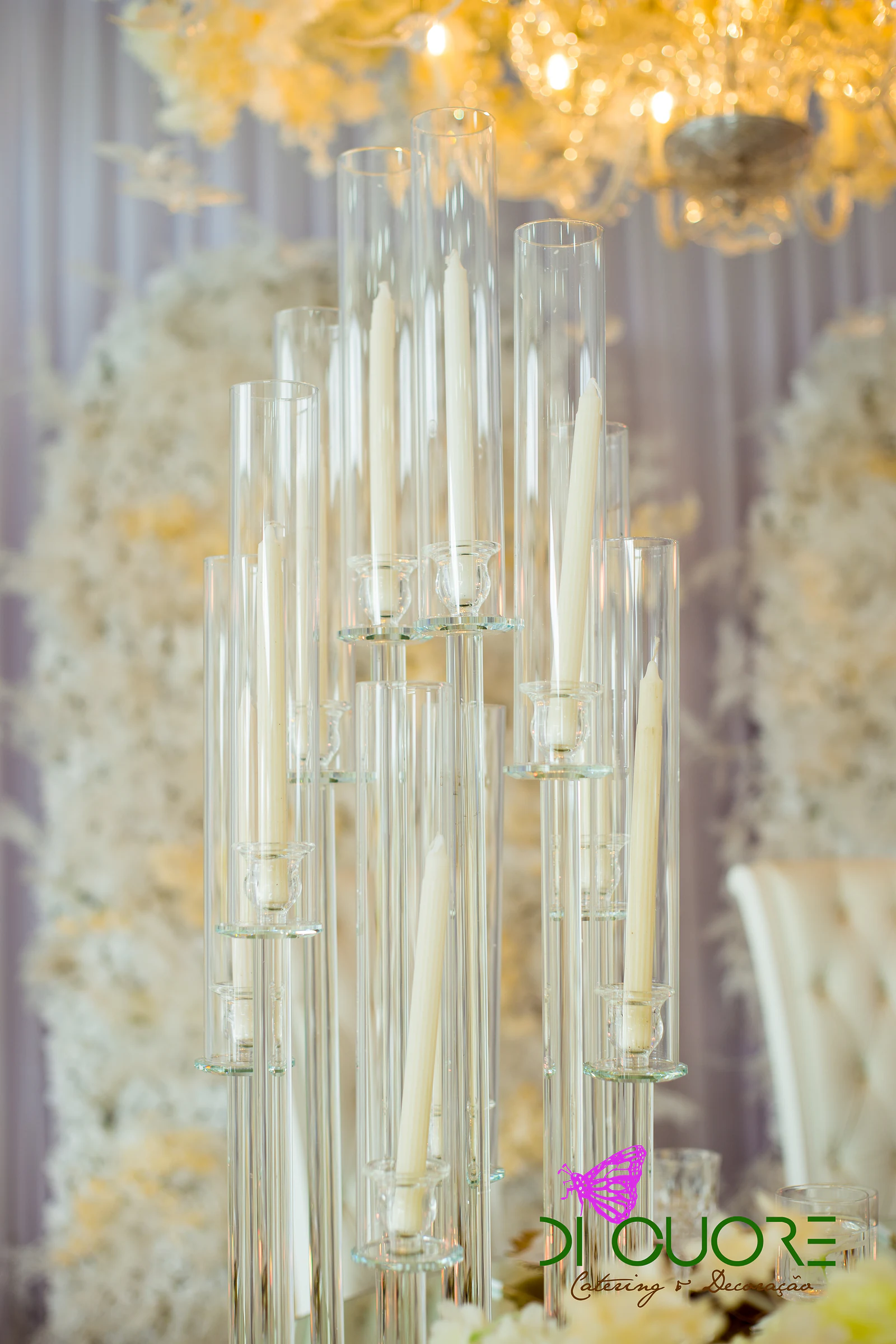 Candelabro de vidro com velas brancas diante de uma parede de flores creme e de um cortinado lilás