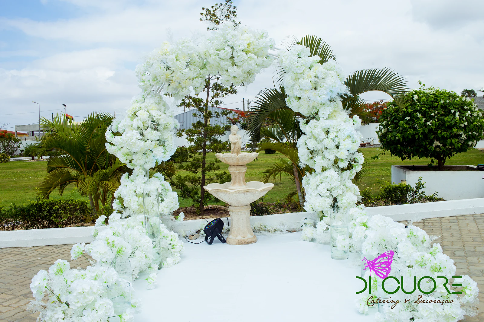 Arco de flores brancas sobre uma passadeira, com uma fonte e um jardim ao fundo