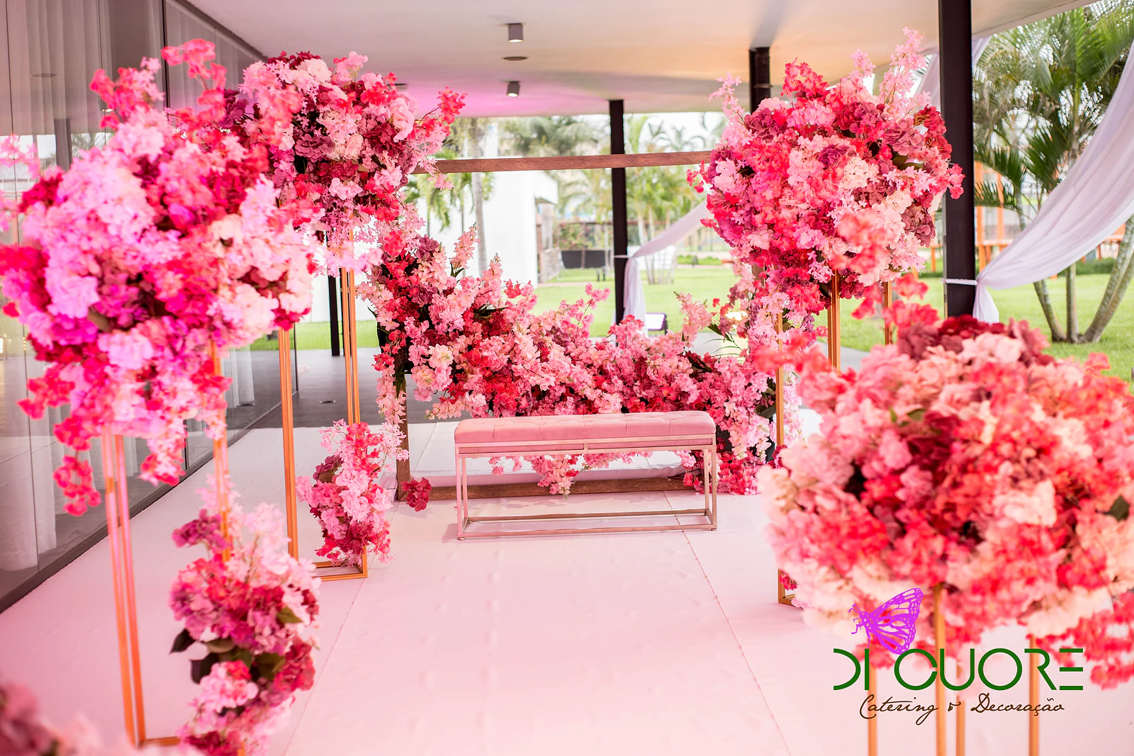 Recanto com banco estofado rosa, arranjos de flores cor-de-rosa em estruturas douradas e vista para o relvado