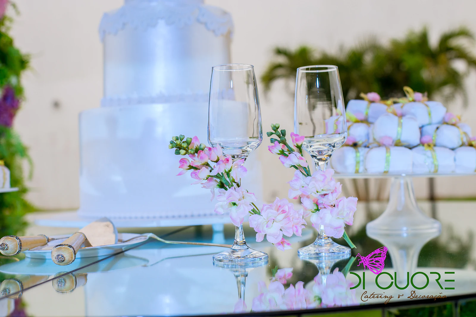 Duas flutes enfeitadas com ramos de flores cor-de-rosa sobre mesa de espelho, com o bolo branco e doces embrulhados ao fundo