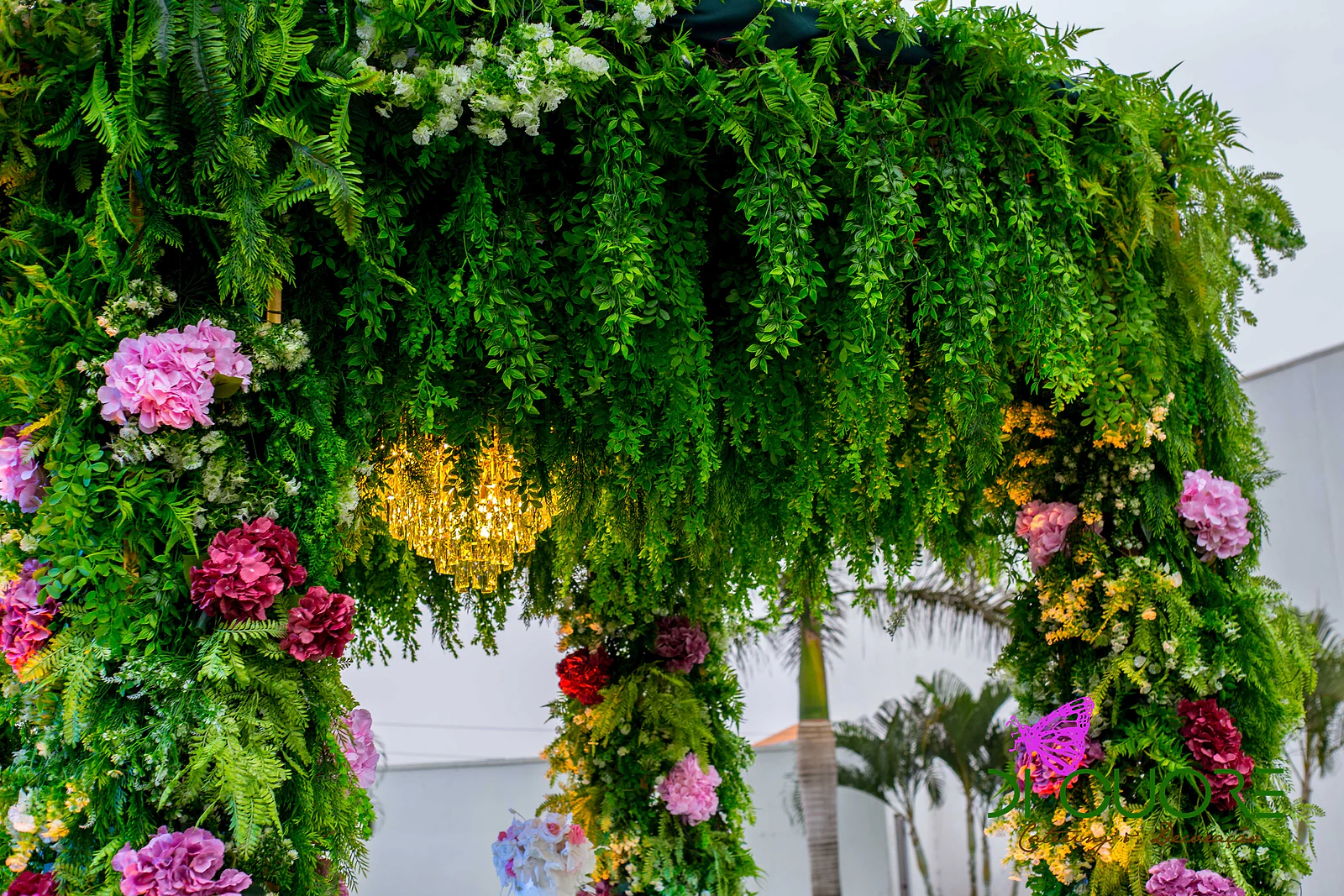 Arco de fetos e flores cor-de-rosa e roxas com um pequeno lustre dourado ao centro