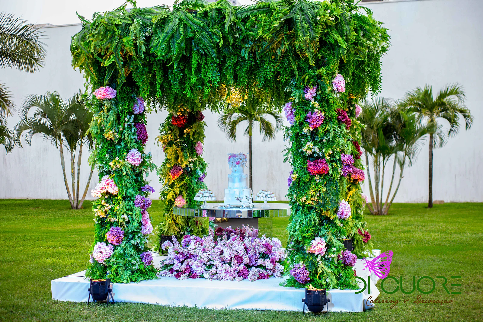 Caramanchão de fetos com hortênsias e flores roxas sobre plataforma branca no relvado, com o bolo numa mesa de espelho ao centro