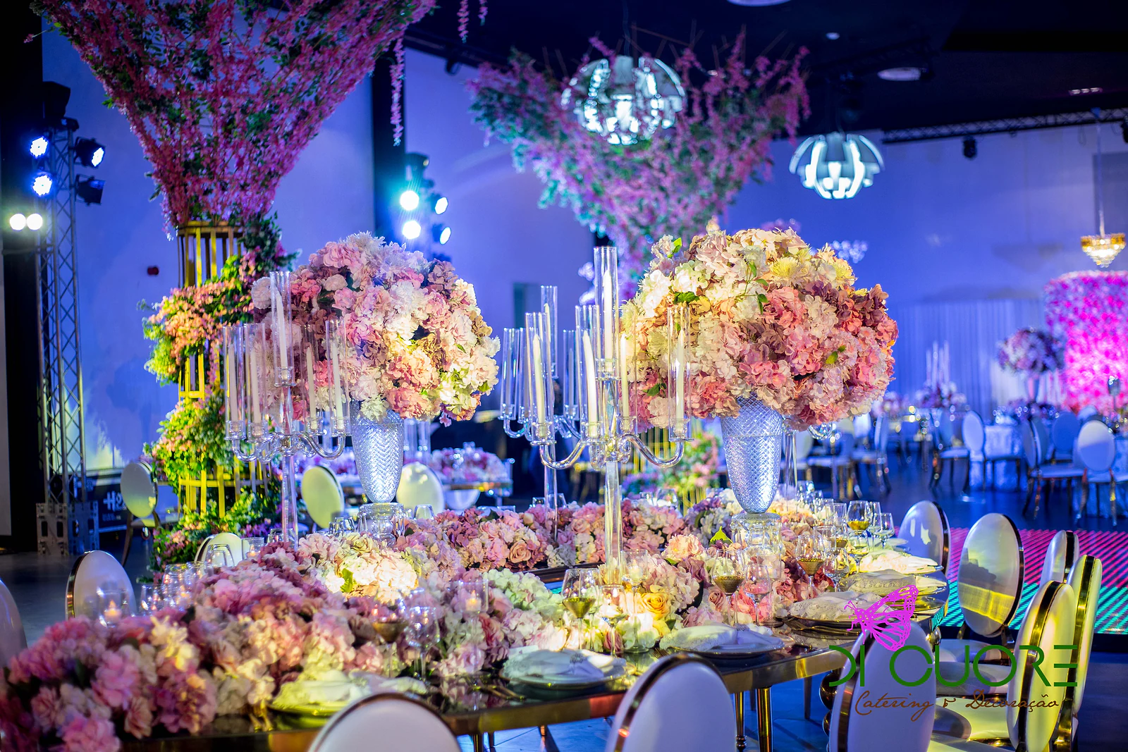Mesa de espelho com candelabros de cristal e esferas de flores em tons de rosa e pêssego, com árvores floridas ao fundo