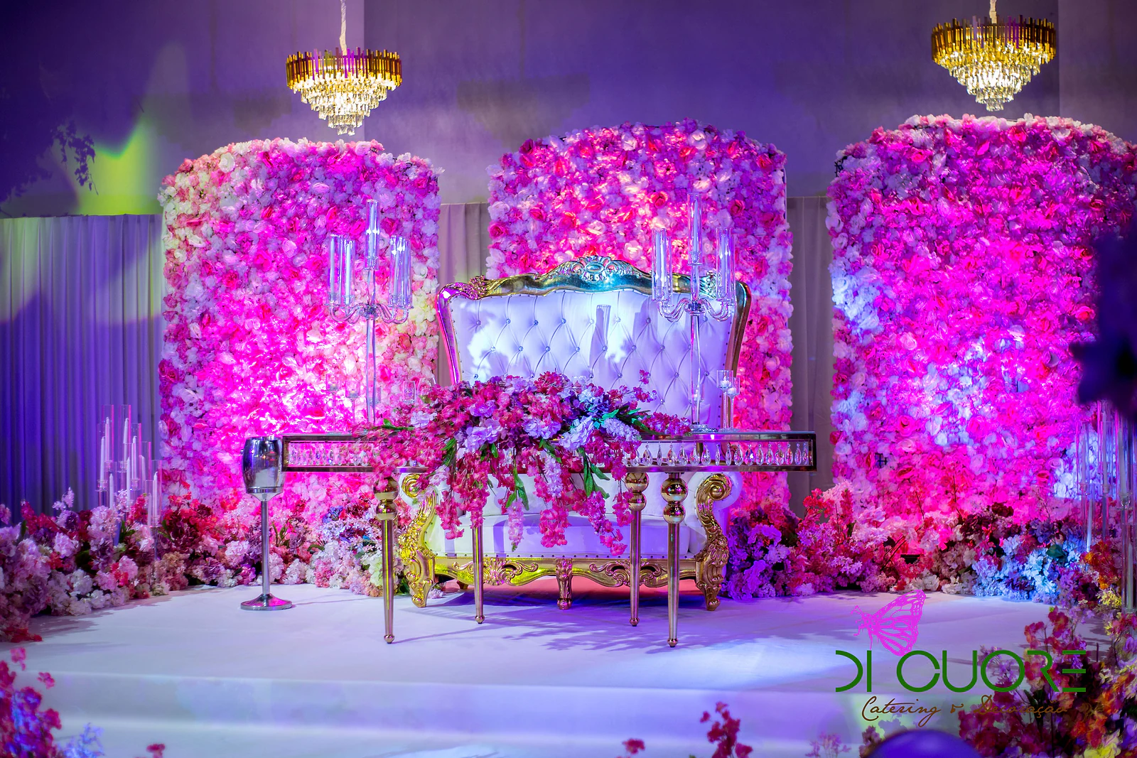 White and gold tufted sofa in front of panels of pink and fuchsia flowers, with a golden chandelier and a floral arrangement on the table in front, in purple light