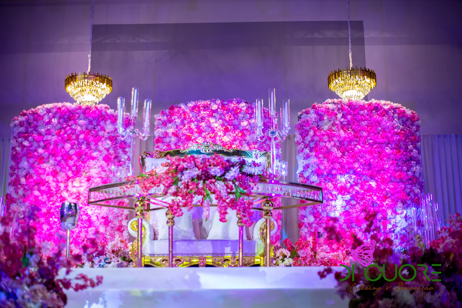 Palco com três colunas de flores fúcsia e lilás, lustres dourados e um sofá branco com arranjo de flores cor-de-rosa sobre estrutura dourada