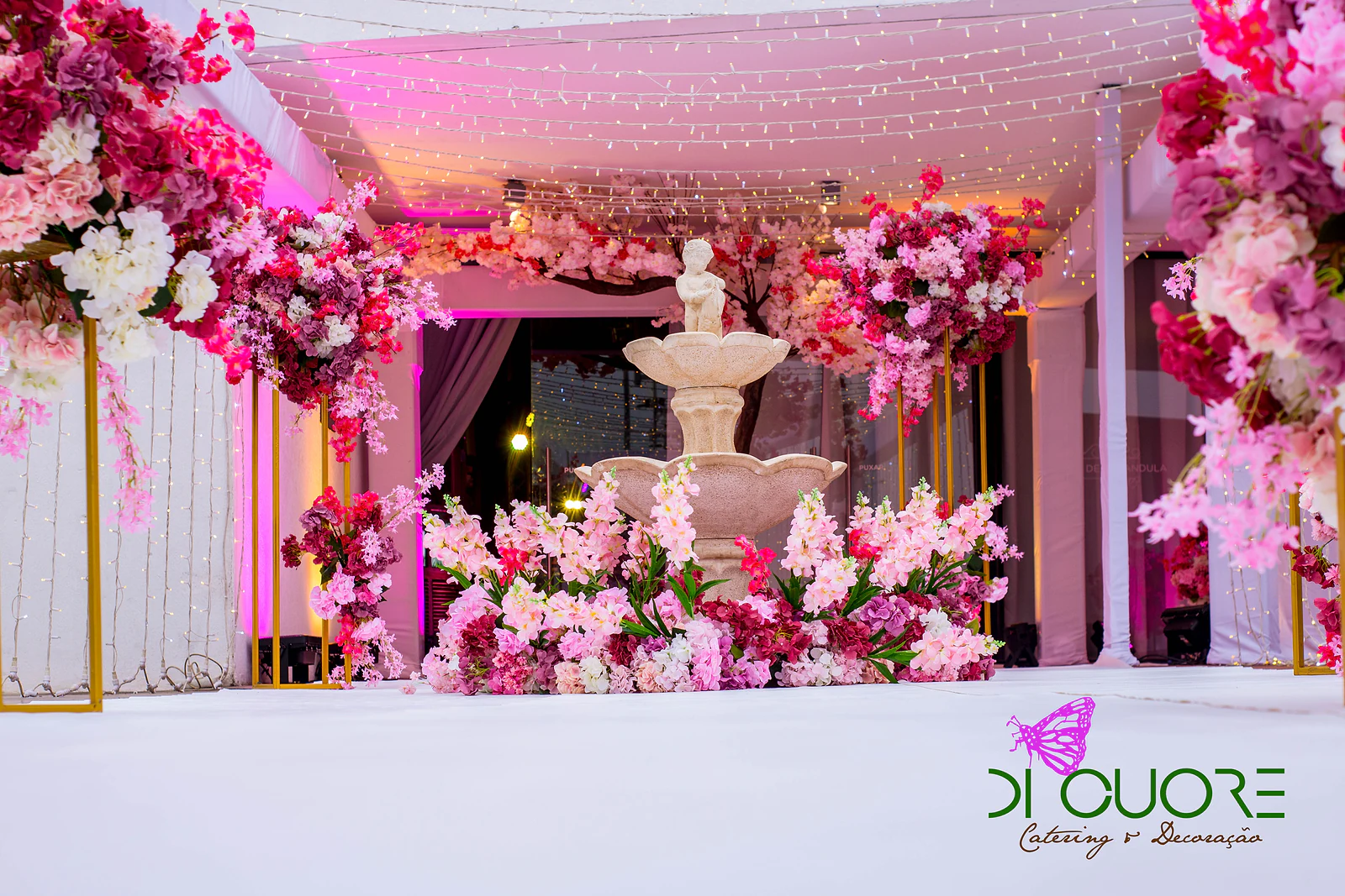 Pérgula branca com cordões de luzes, fonte rodeada de flores cor-de-rosa e vasos brancos com arranjos fúcsia