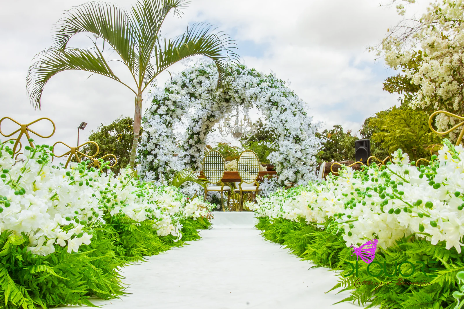 Corredor branco ladeado de flores brancas e fetos, em direcção ao arco de flores, com uma palmeira e laços dourados nas cadeiras