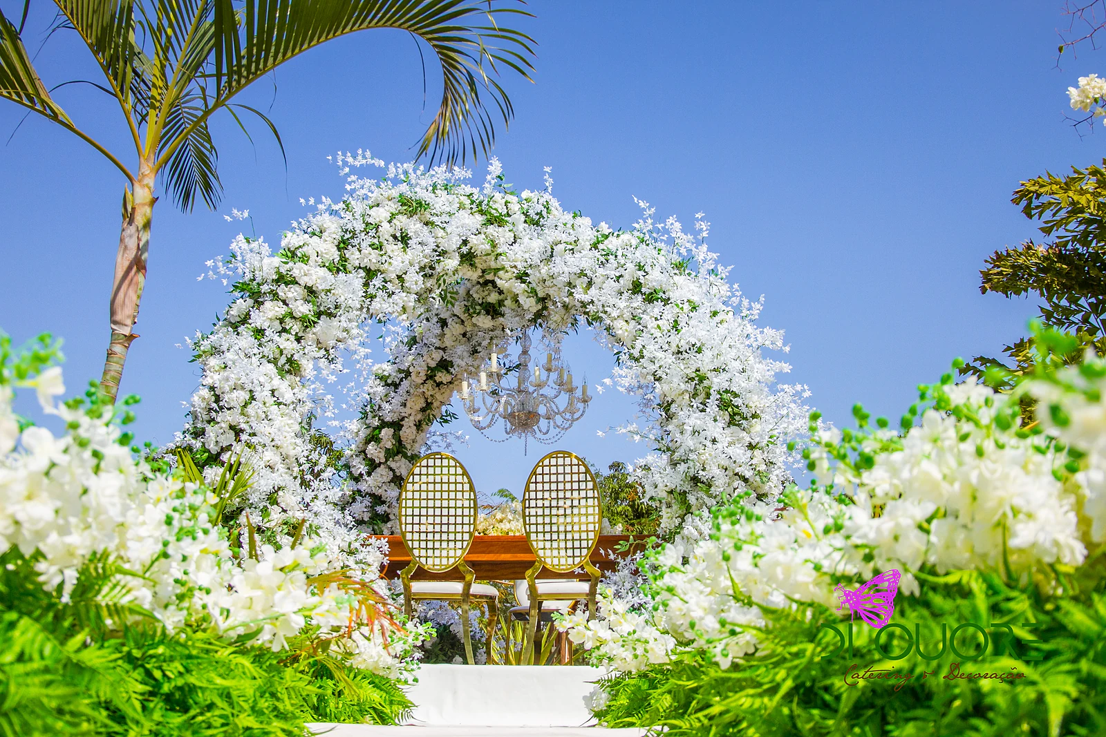 Arco redondo de flores brancas com um lustre de cristal e duas cadeiras douradas ao fundo de um corredor branco ladeado de fetos e flores, sob um céu azul e uma palmeira