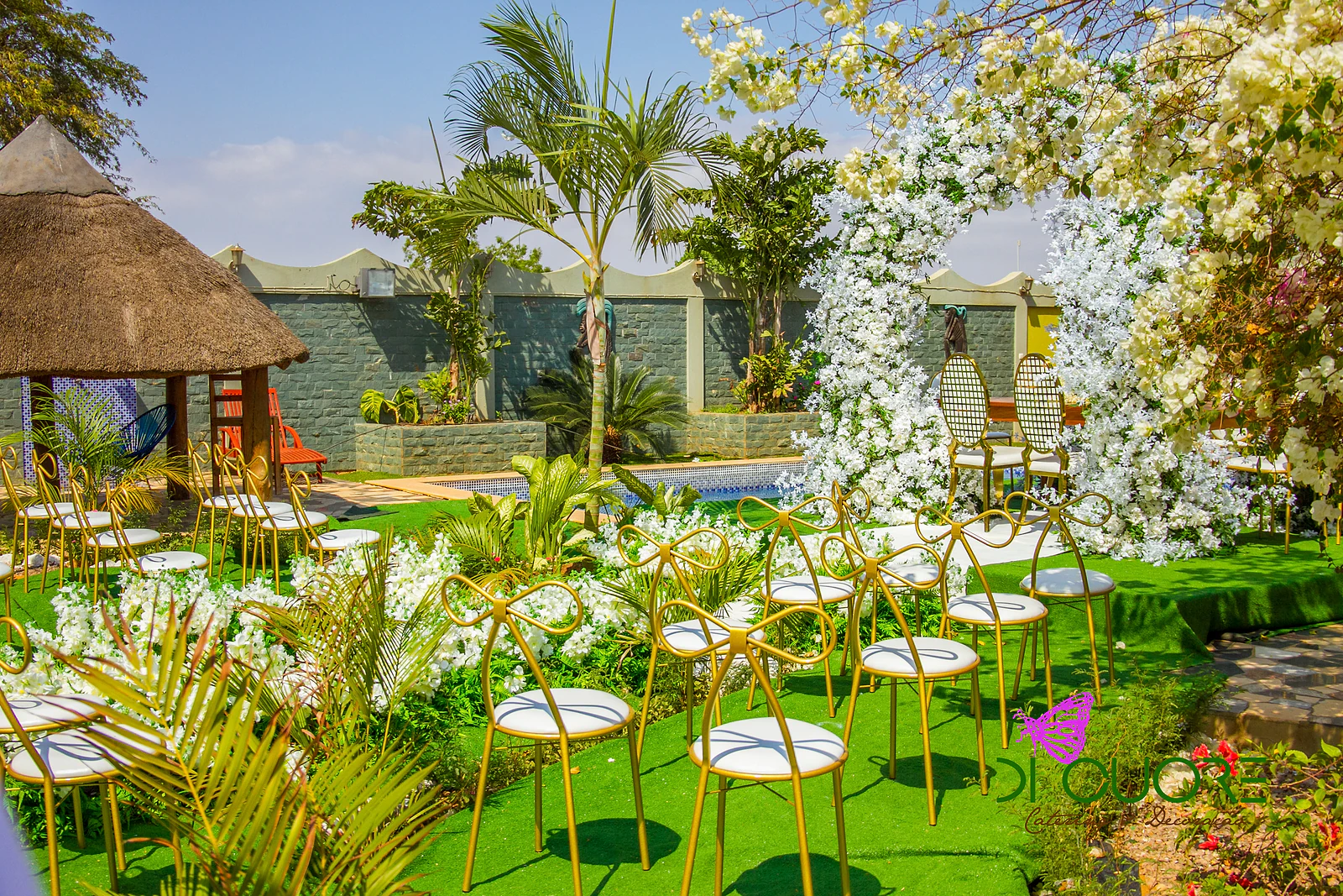 Cadeiras douradas com laço e assento branco sobre relva junto à piscina, com o arco de flores brancas ao fundo e uma palmeira