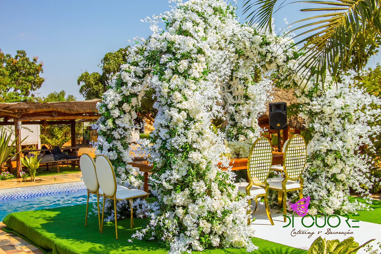 Arco de flores brancas visto de lado, com a piscina à esquerda, duas cadeiras douradas de espaldar oval e uma palmeira à direita