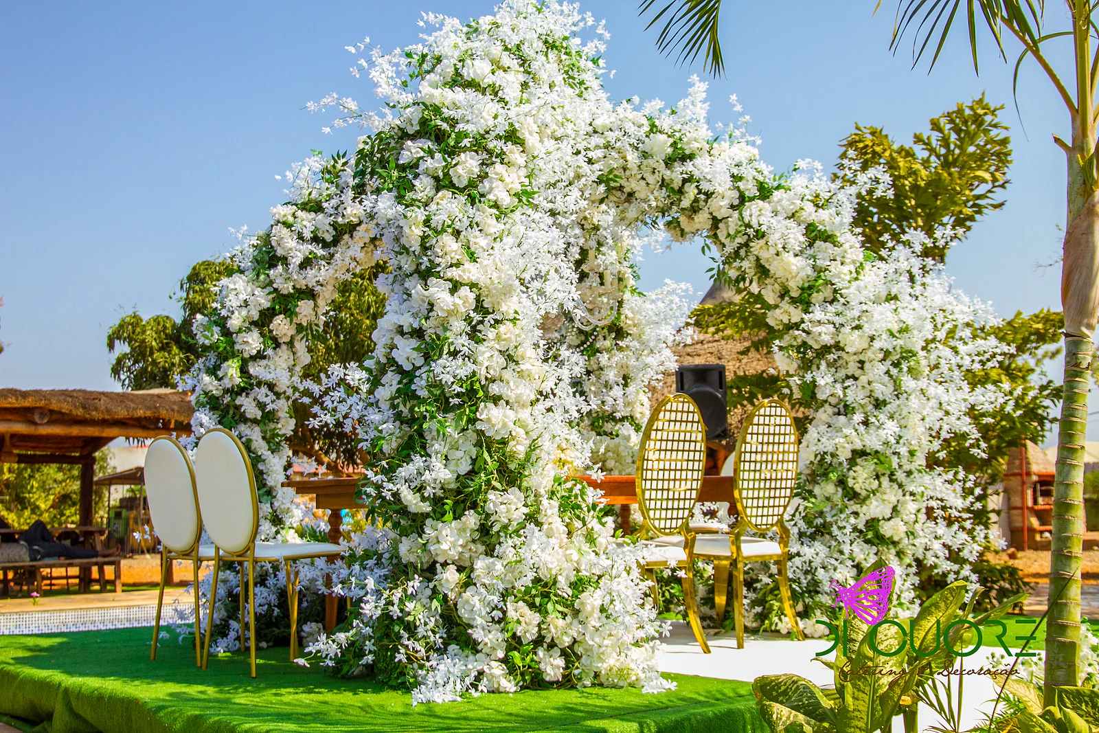 Arco de flores brancas visto de lado, com cadeiras douradas e brancas, uma palmeira e um céu azul