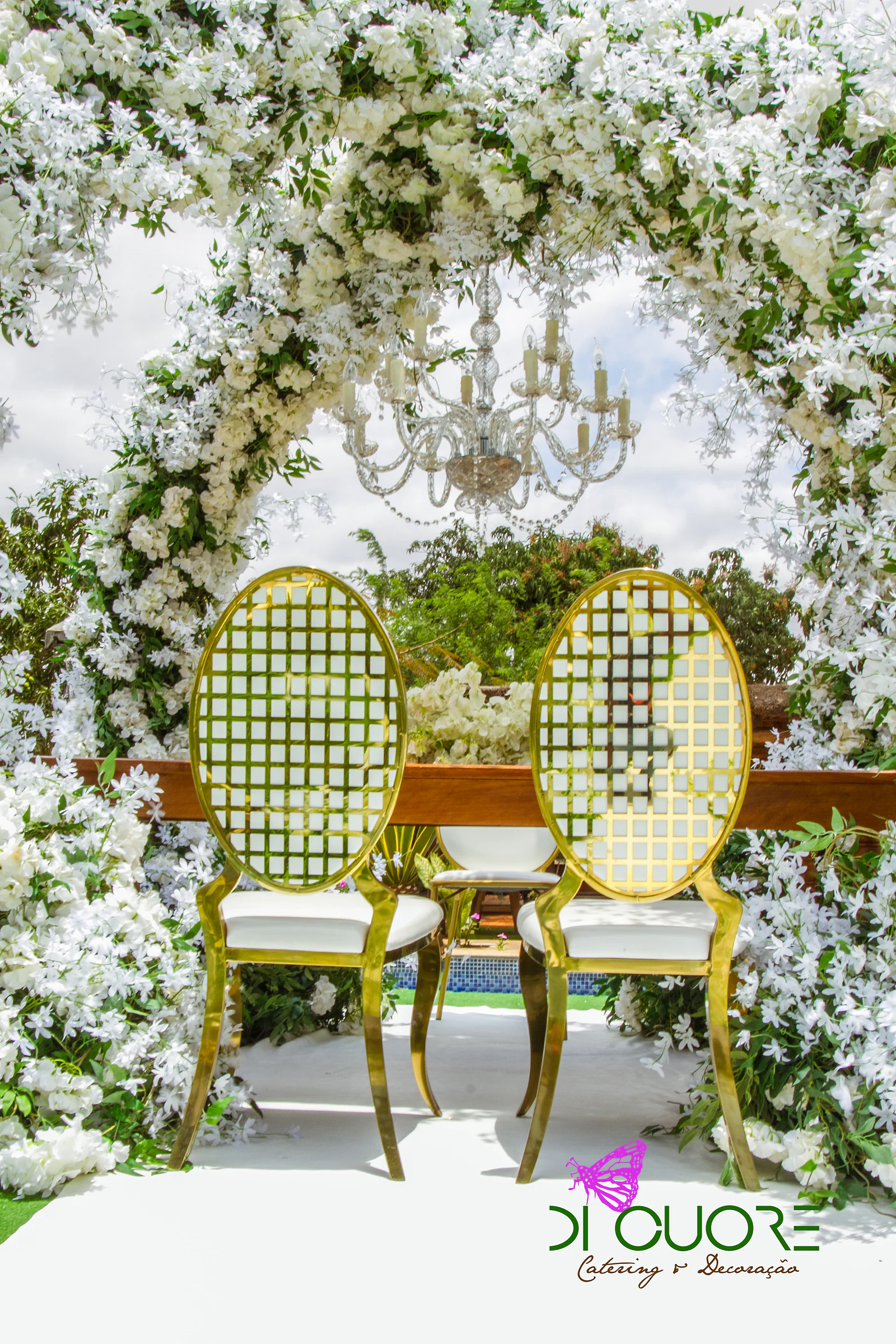 Duas cadeiras douradas de espaldar em treliça e assento branco sob o arco de flores brancas e um lustre de cristal