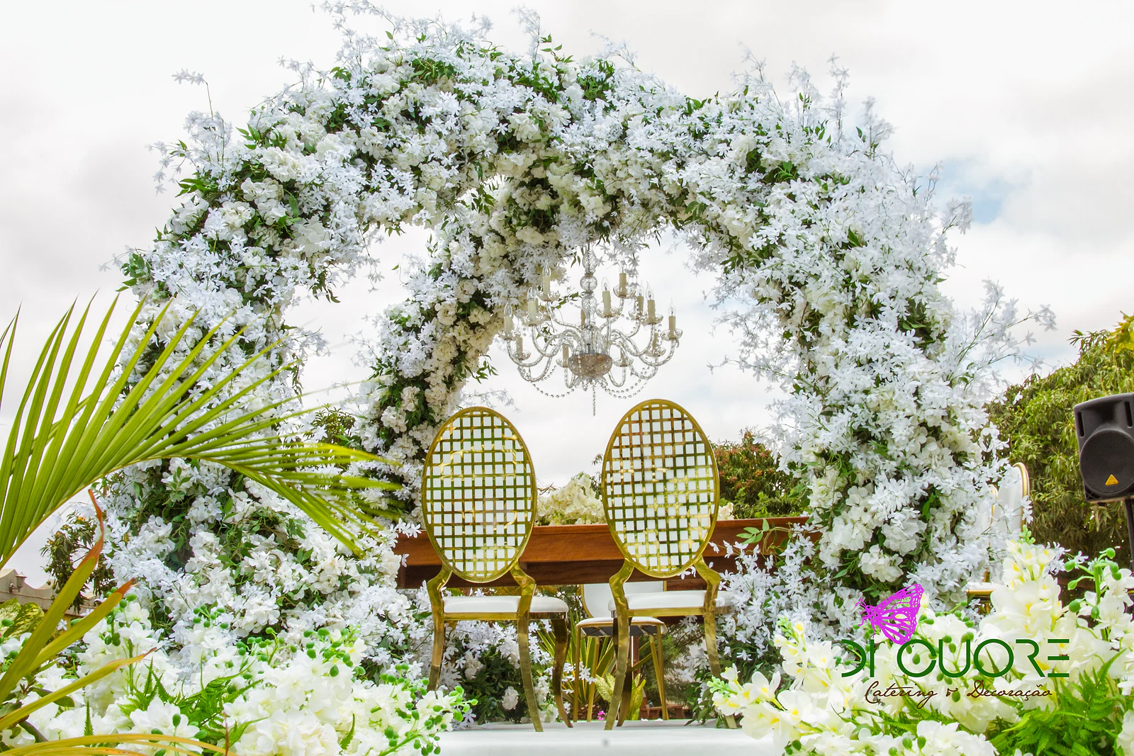 Arco redondo de flores brancas e folhagem, com um lustre de cristal suspenso no interior e duas cadeiras douradas de espaldar oval em treliça diante de uma mesa de madeira