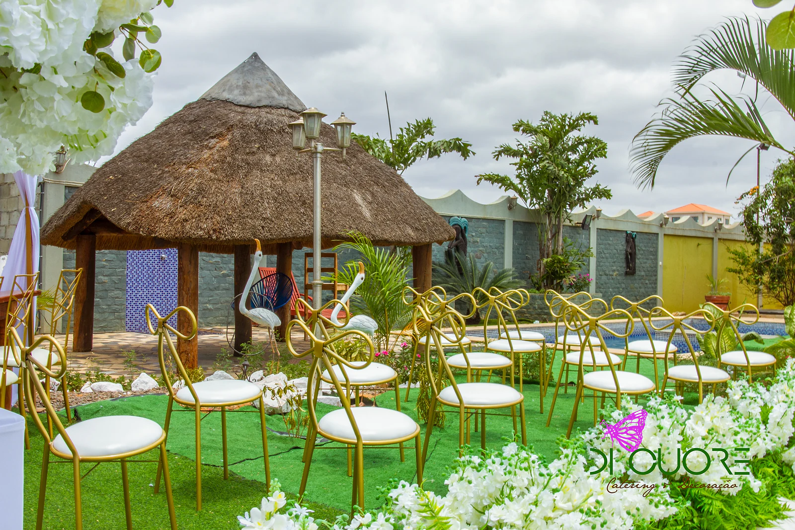 Cabana de palha com candeeiro de jardim e cadeiras douradas com laço sobre relva, junto à piscina, com flores brancas em primeiro plano
