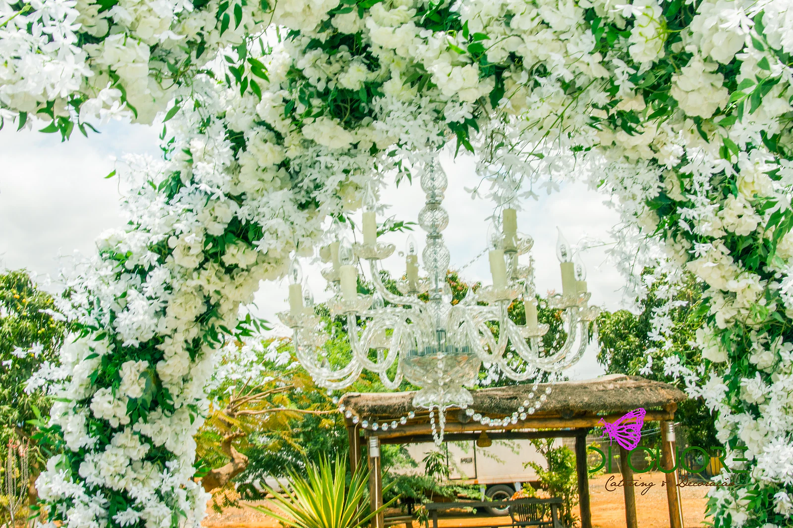 Lustre de cristal com velas suspenso dentro do arco de flores brancas e folhas verdes, com uma cabana de palha ao fundo