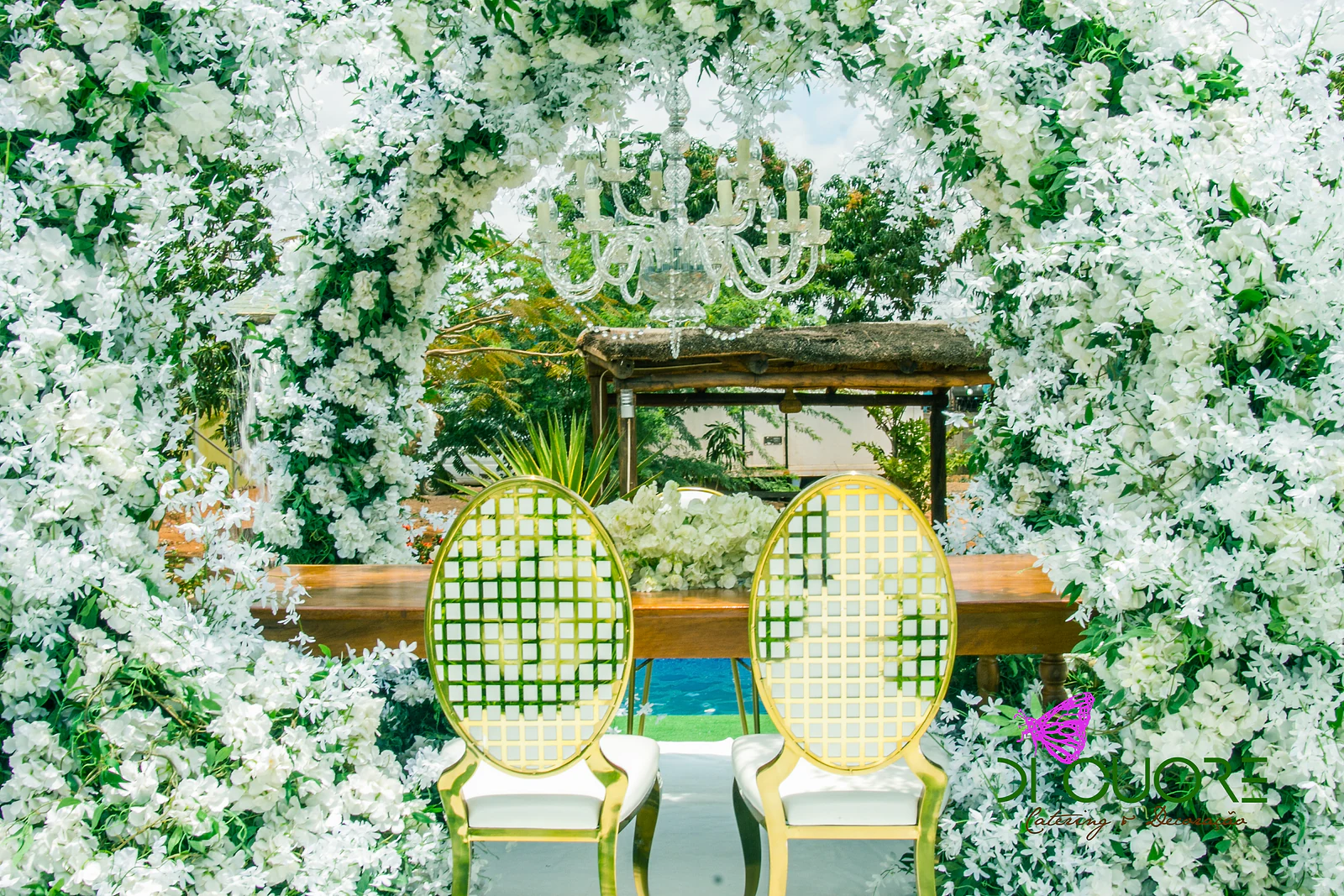 Duas cadeiras douradas de espaldar em treliça diante de uma mesa de madeira, emolduradas por flores brancas e folhas verdes, com o lustre de cristal por cima