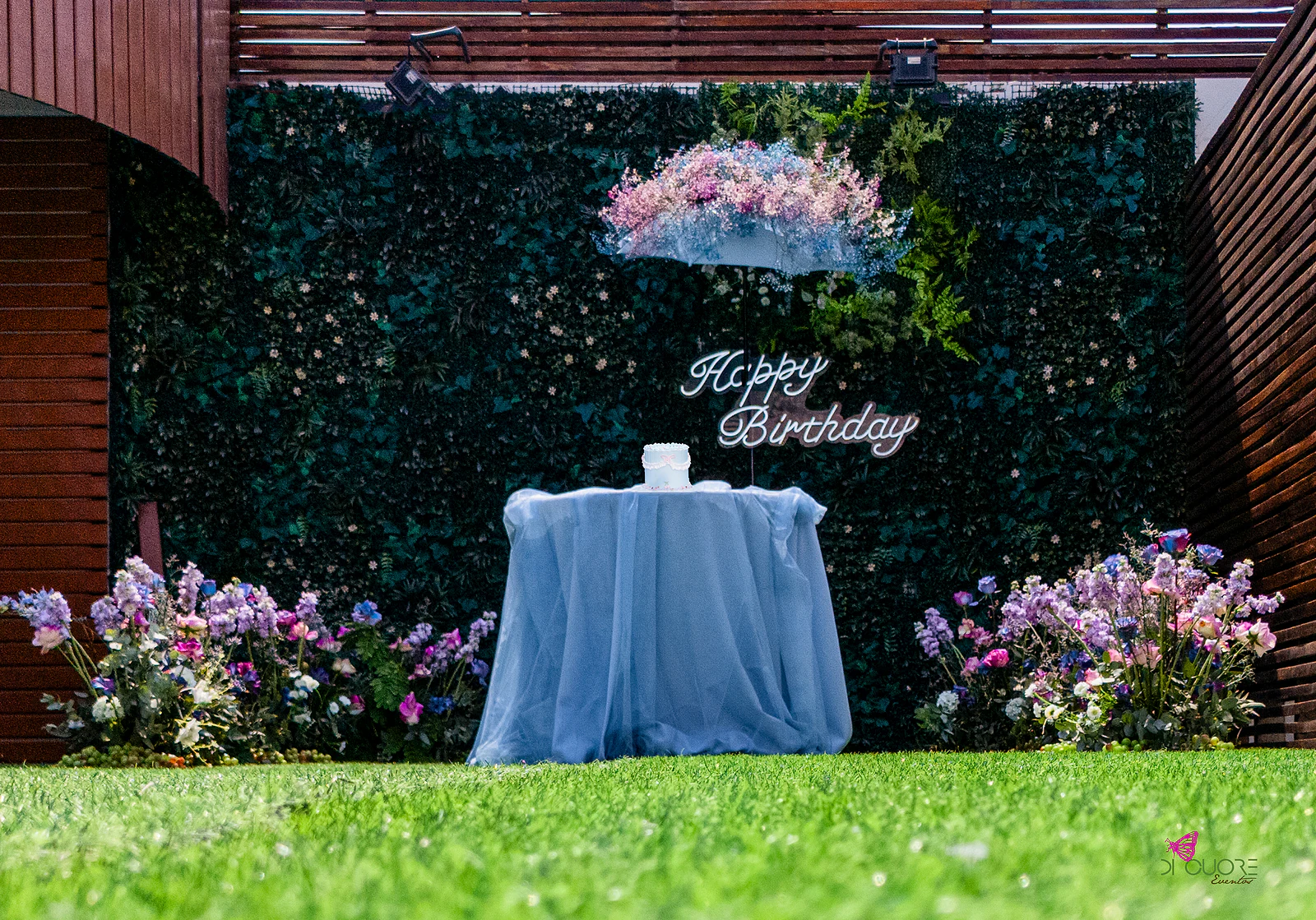 Mesa do bolo com tule azul-claro diante do muro de folhagem, entre dois canteiros de flores lilás e rosa sobre a relva