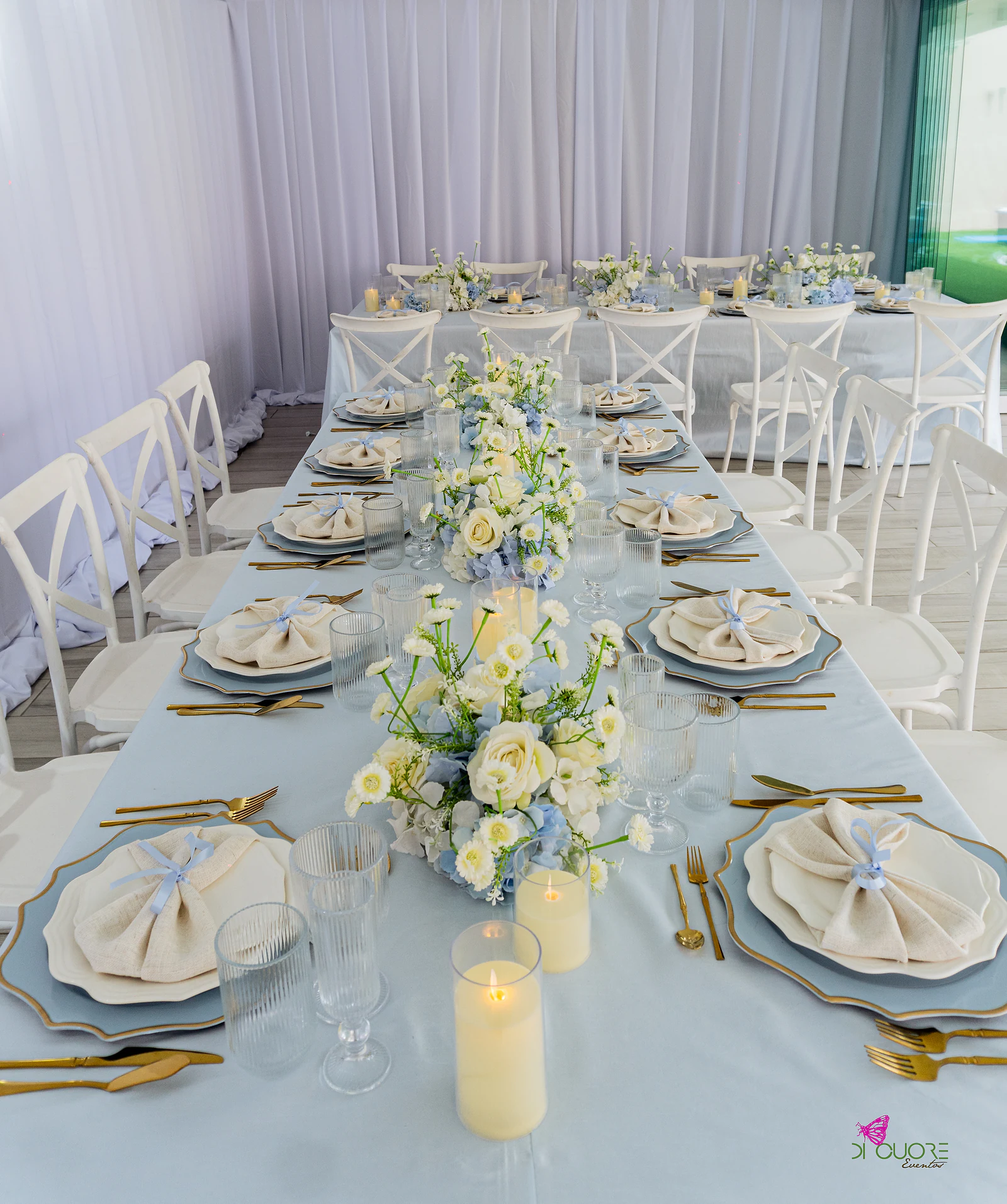 Mesa comprida com toalha azul-claro, pratos azuis com rebordo dourado, guardanapos atados com fita, velas e um ramo de rosas brancas e margaridas