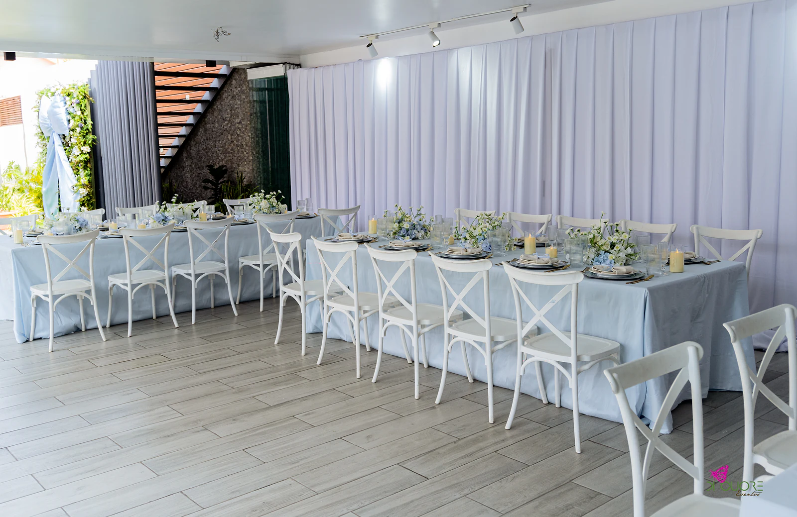 Sala com mesas compridas de toalha azul-claro, cadeiras brancas de costas cruzadas e cortinas brancas ao fundo