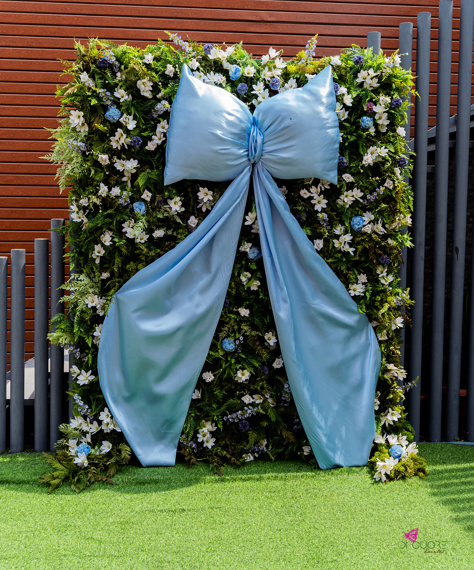Painel de folhagem e flores brancas com um laço gigante de cetim azul-claro, junto a ripas de madeira